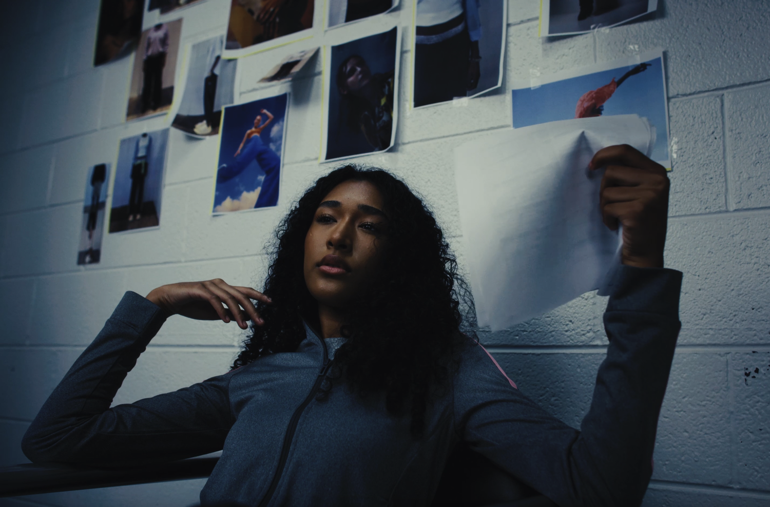 A young woman with curly black hair sitting against a white brick wall filled with colorful photographs of athletes and models, holding a crumpled sheet of paper in her right hand, and resting her left hand near her face.