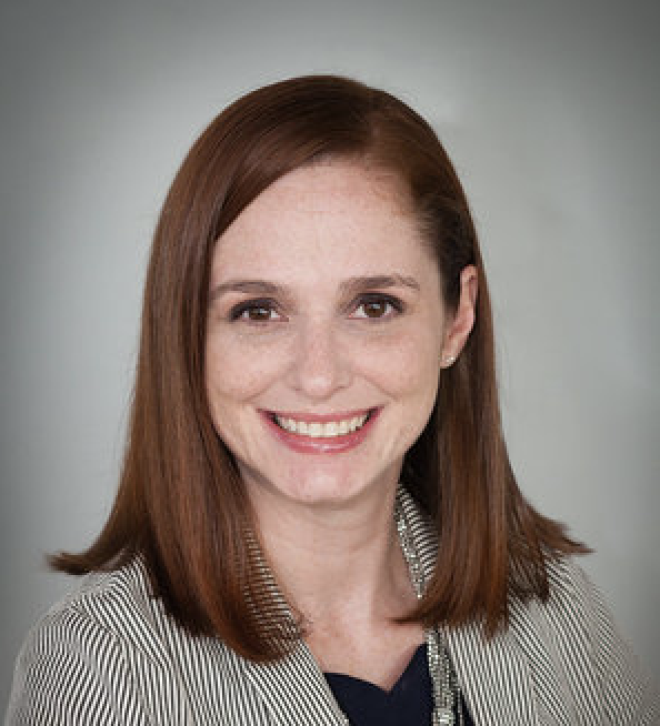 A woman with shoulder-length auburn hair smiling at the camera, wearing a black top and a patterned blazer, against a gray background.