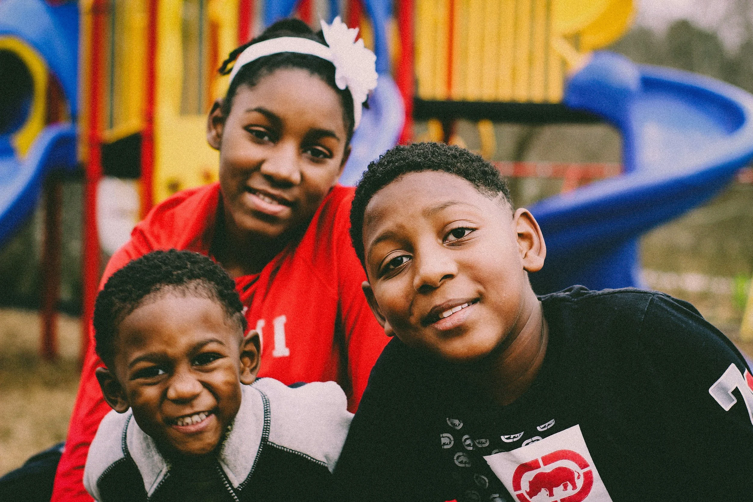 A group of smiling children and a woman at an outdoor event with colorful playground equipment in the background.
