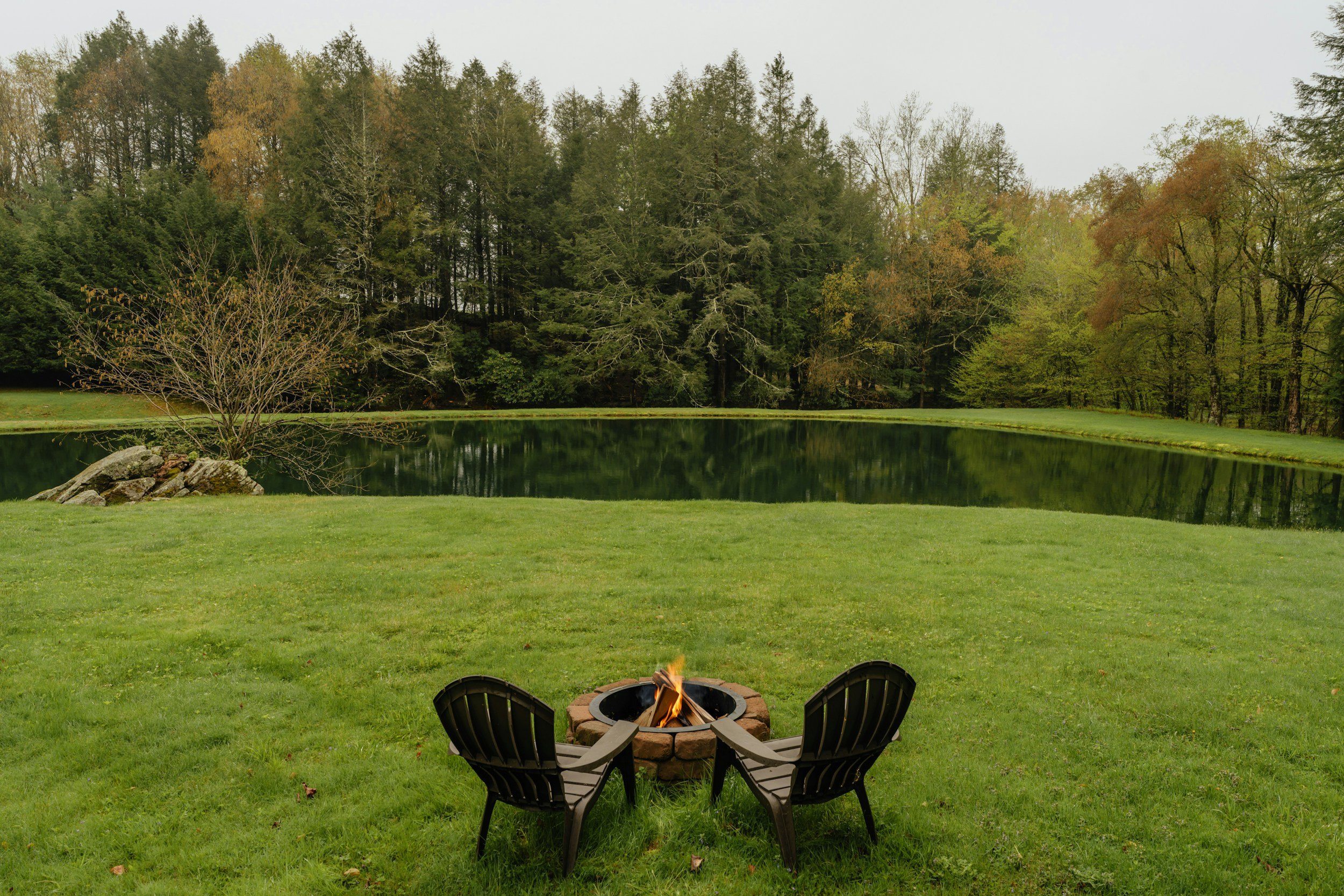 A backyard scene with a fire pit surrounded by three chairs, overlooking a small lake with trees in the background. The sky is overcast.