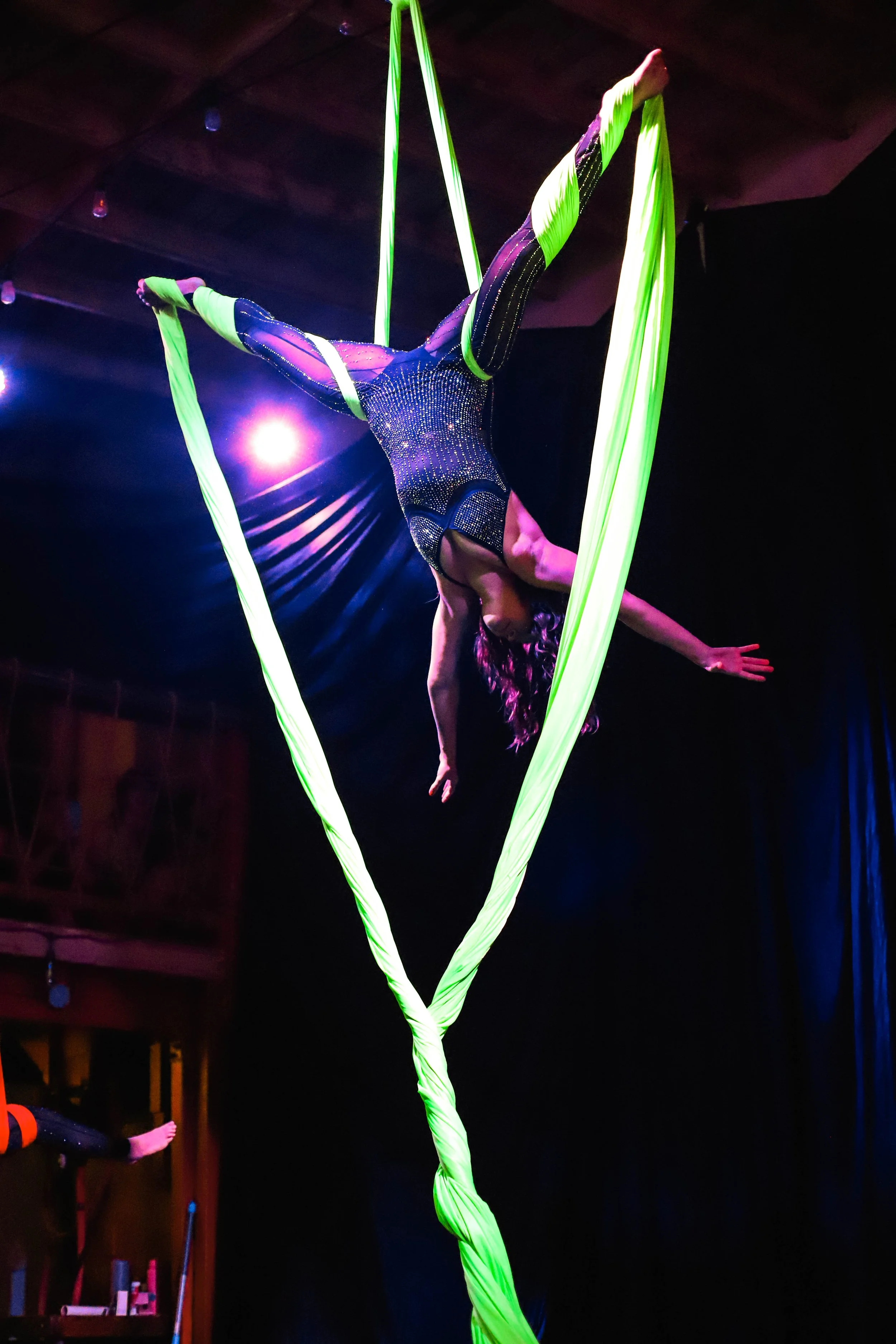 Aerial silk performer in a black and green costume performing on bright green fabric in a dark setting with stage lights.