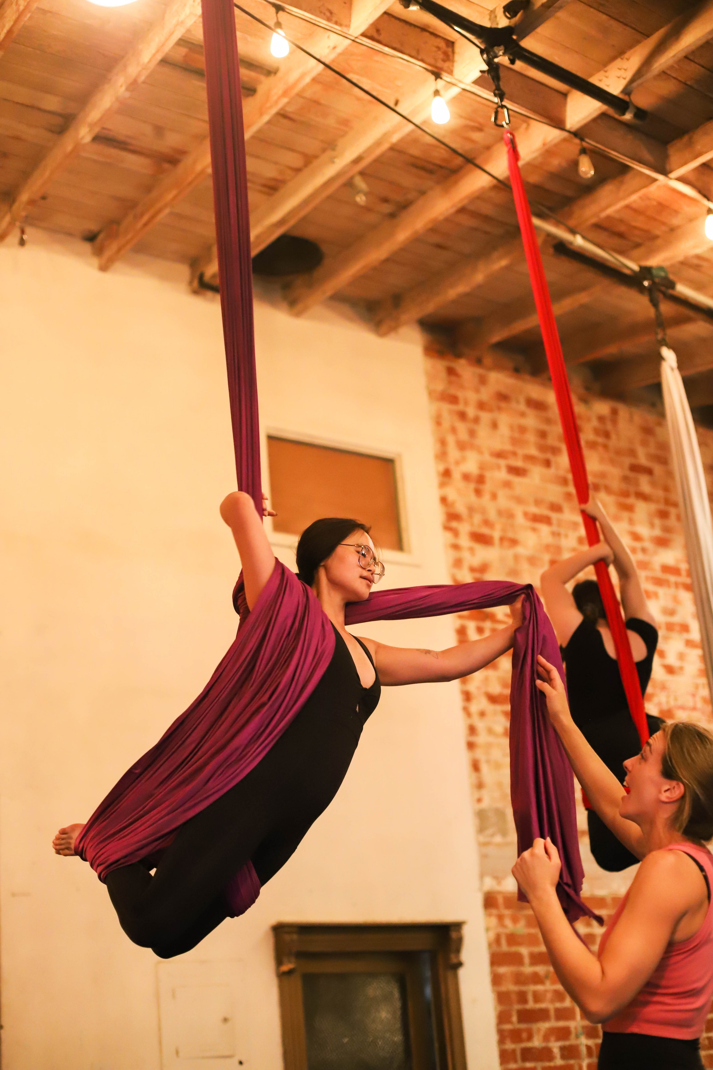 Two women practicing aerial silk acrobatics indoors, with one woman in black clothing suspended on purple silks and another woman in pink top holding the silks, in a room with a brick wall and wood ceiling.
