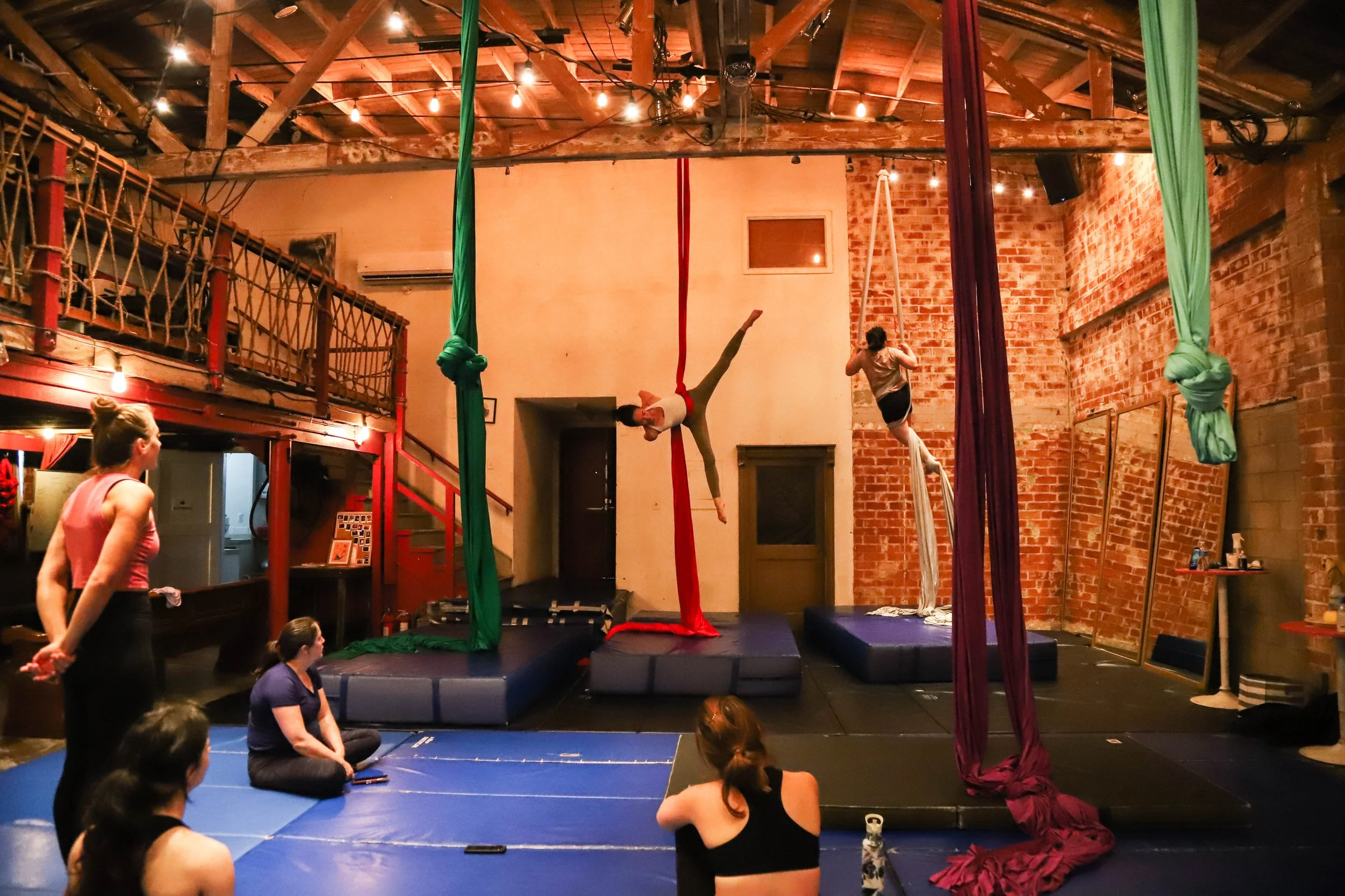 Aerial silk performers practicing in a gym with brick and wooden interior. One performer is hanging upside down on green silk, another on red silk, and a third on purple silk. Several people sit on the floor watching, and others are standing or seated nearby.