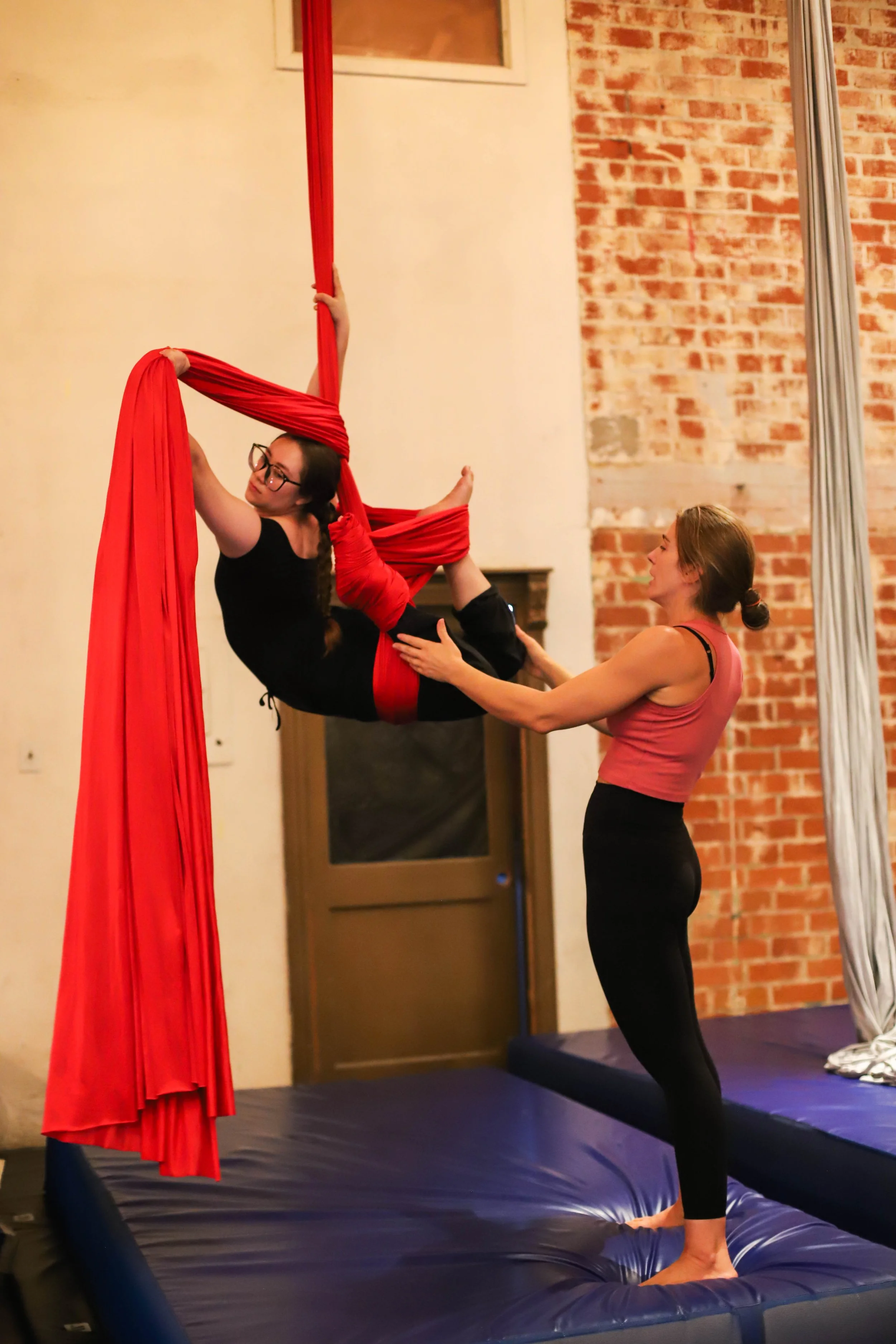 Two women practicing aerial silk therapy indoors on a blue trampoline with a brick wall in the background. One woman is upside down, holding onto red silk fabric with her legs and arms, while the other woman supports her from below.