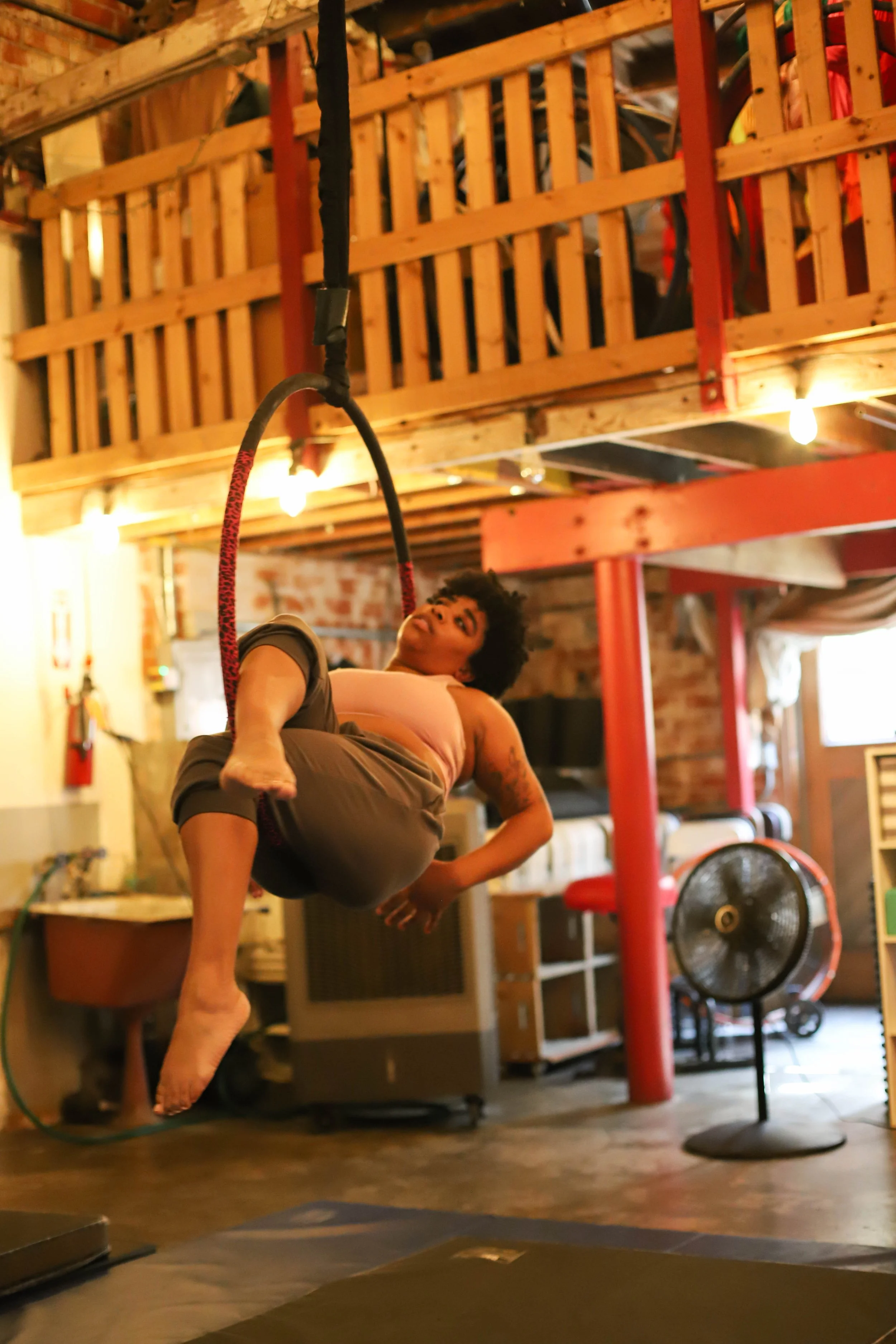 A person practicing aerial silk yoga in a cozy, rustic indoor space with warm lighting, wooden beams, and a fan in the background.