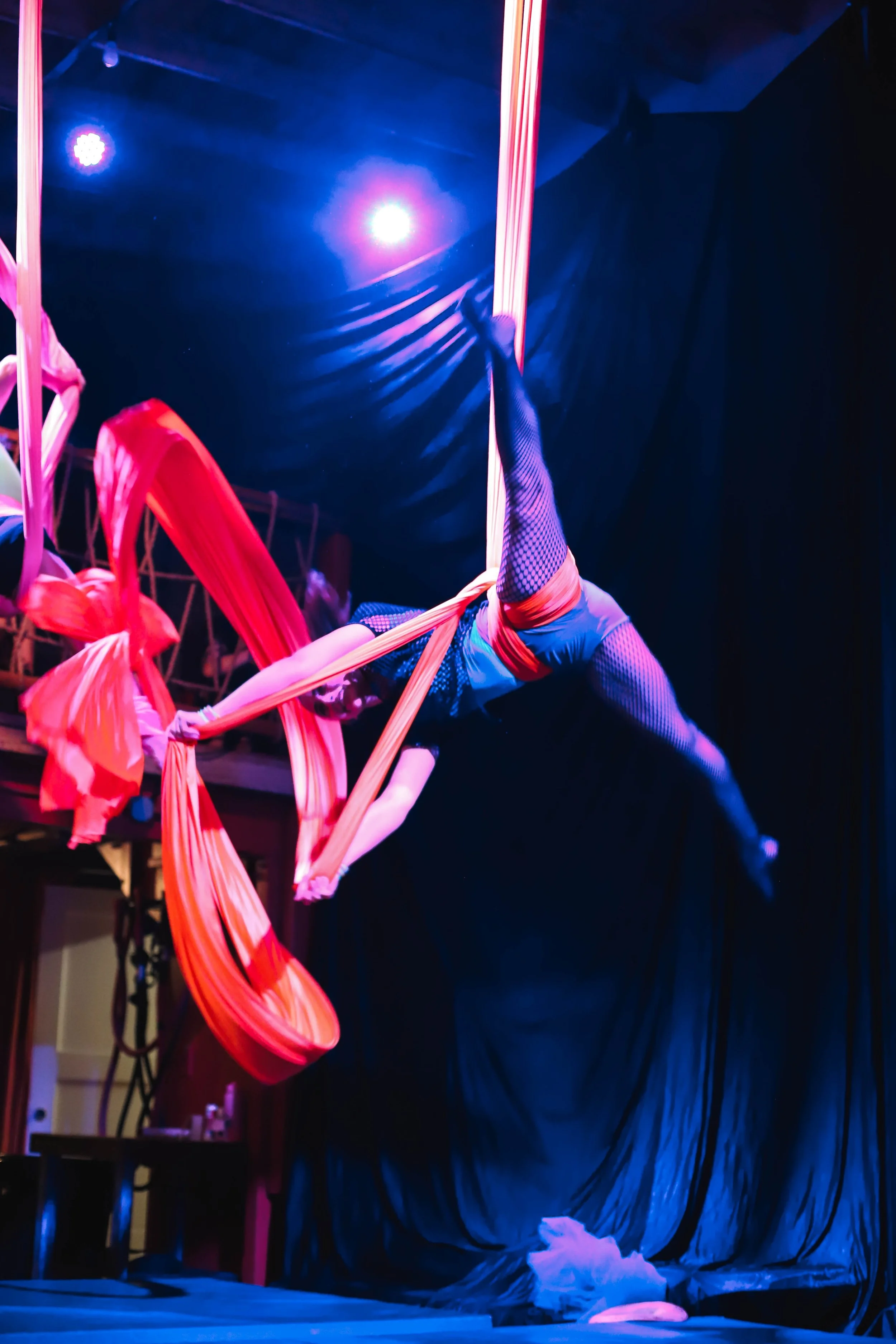 A performer in a blue costume with fishnet tights is hanging upside down from pink fabric ribbons, holding onto them with one hand, on a stage with blue lighting and a dark backdrop.