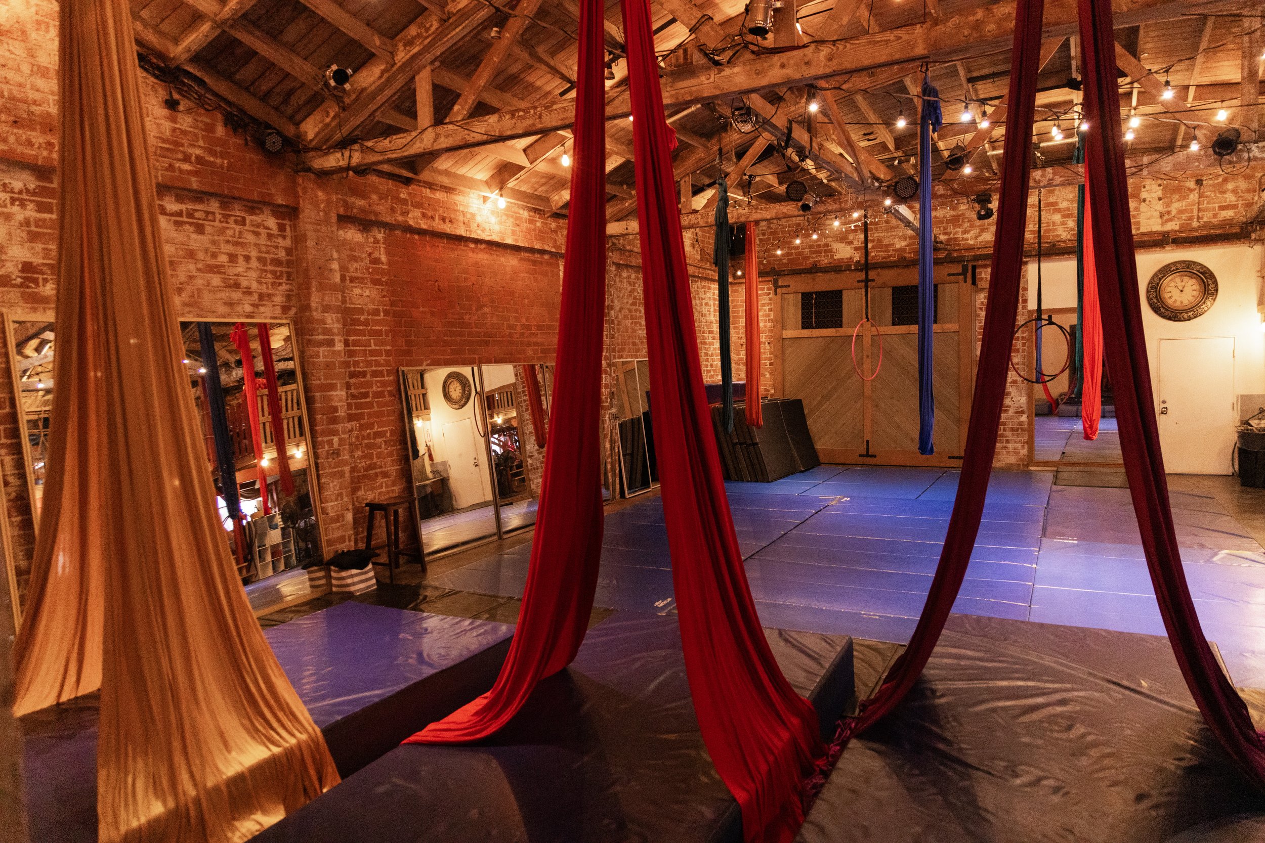 Indoor aerial silk performing area with hanging red, orange, blue, and teal silks, exposed brick walls, string lights on the ceiling, mirrors on the wall, and a clock.