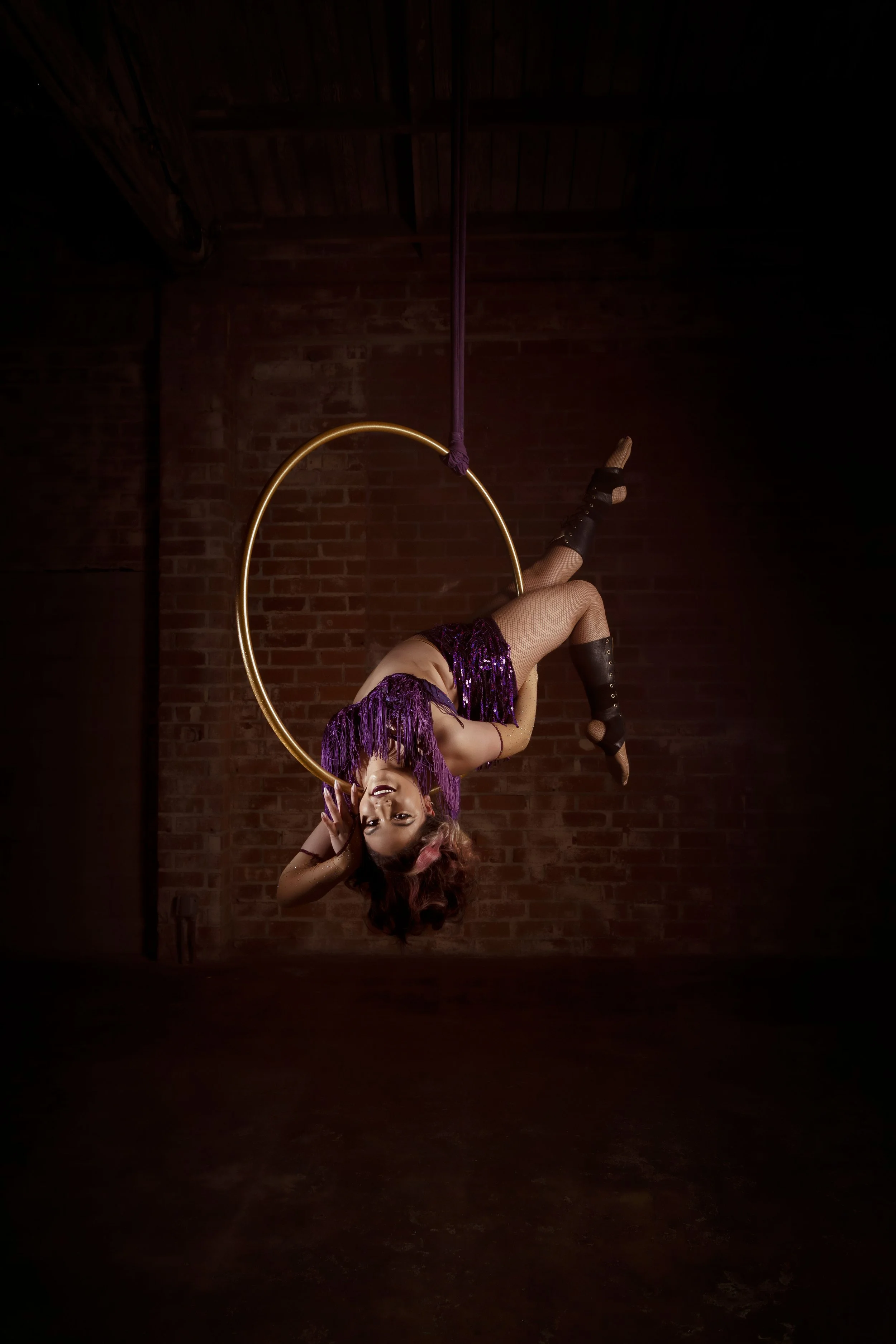 A woman performing aerial silk acrobatics on a purple silk hanging from the ceiling, with a circular aerial hoop nearby, in a dimly lit brick-walled studio.