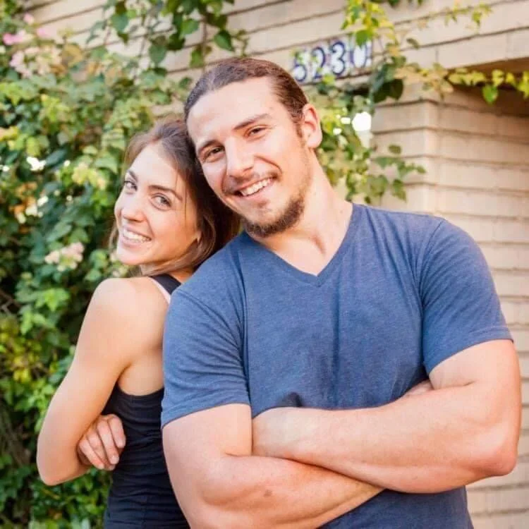 A smiling man and woman leaning back to back outdoors in front of a brick wall and greenery.