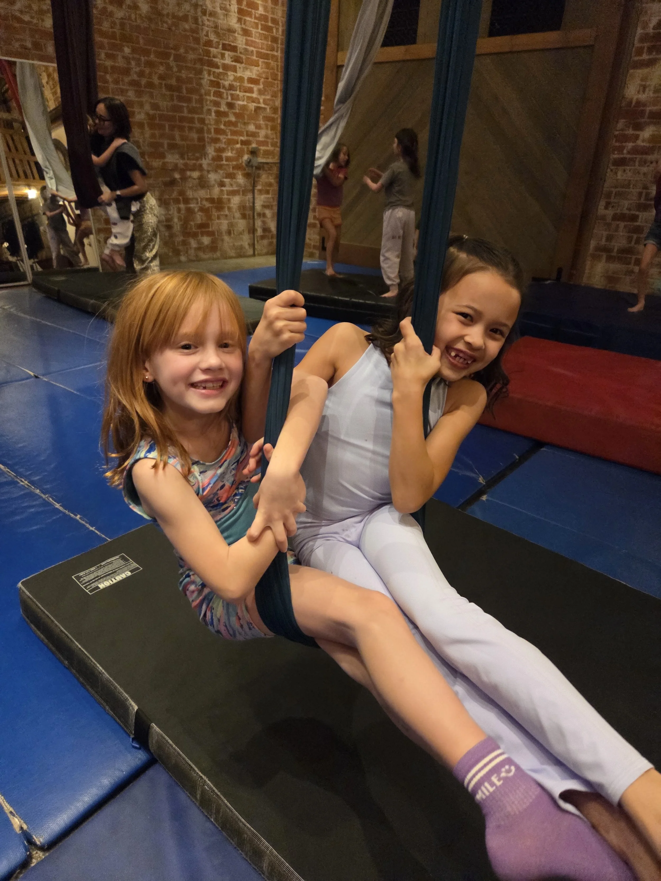 Two young girls smiling and posing on a trampoline at an indoor trampoline park, with other children jumping and playing in the background.