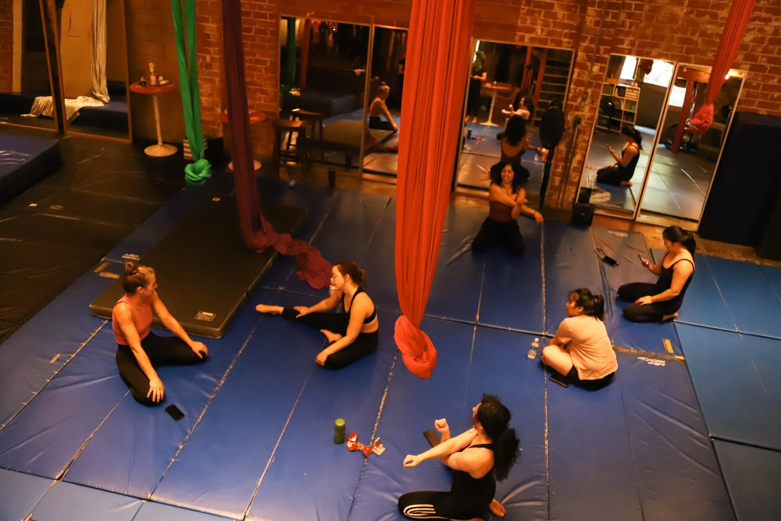 People in an aerial arts studio practicing aerial silks and stretching on mats.
