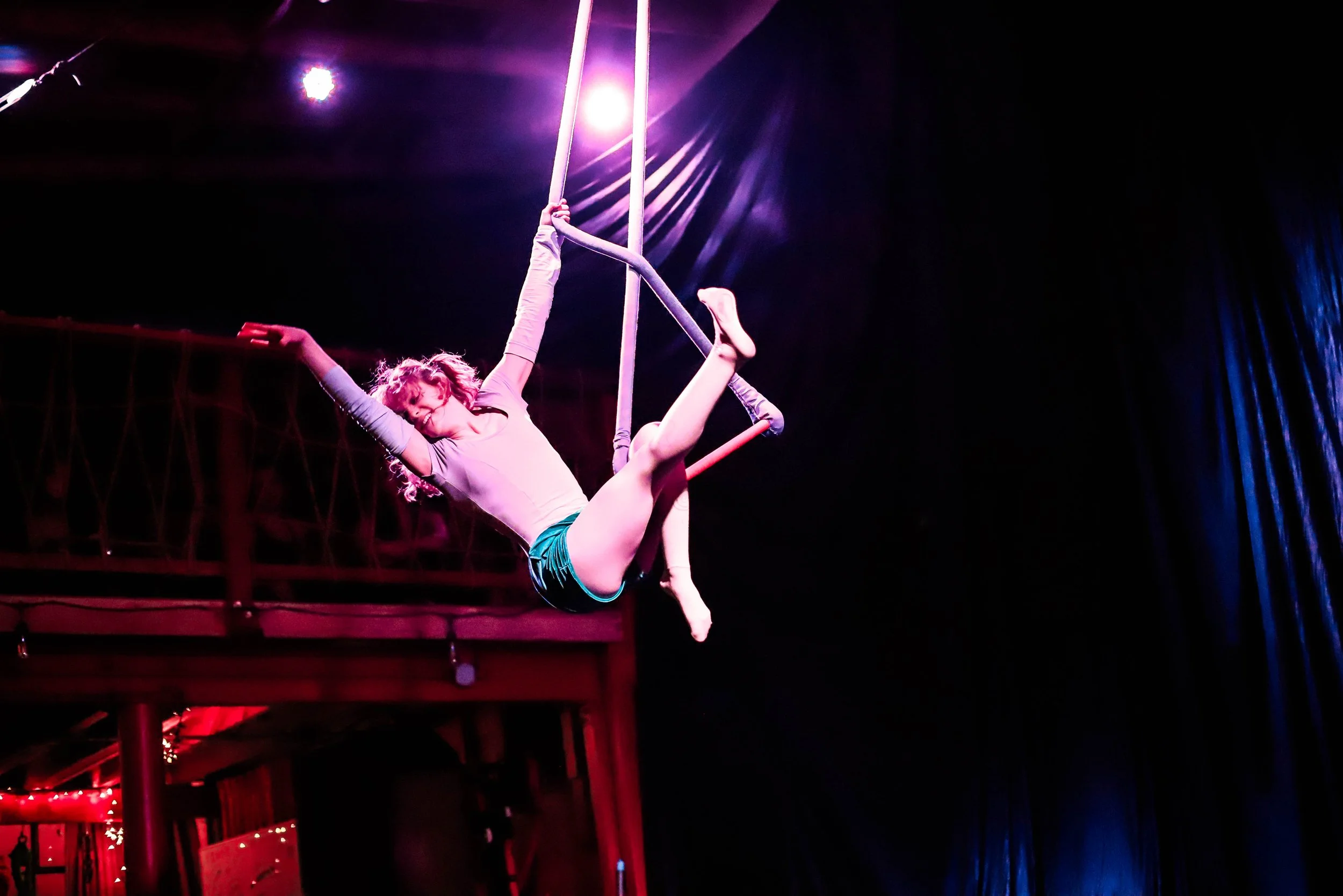 A female aerialist performs on a trapeze with purple stage lighting in the background.