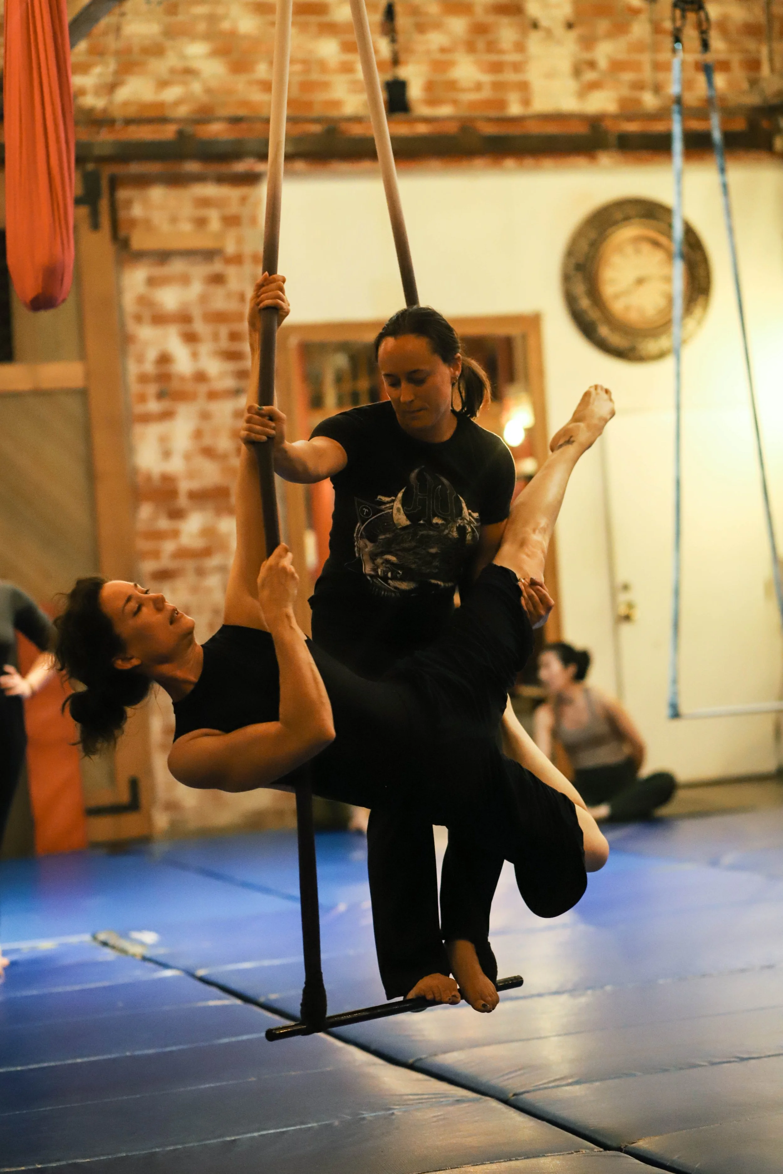 Two women practicing acro yoga with a silk hammock in a gym with exposed brick walls. One woman is holding the suspended woman, who is seated on a pole with her legs extended and head down.