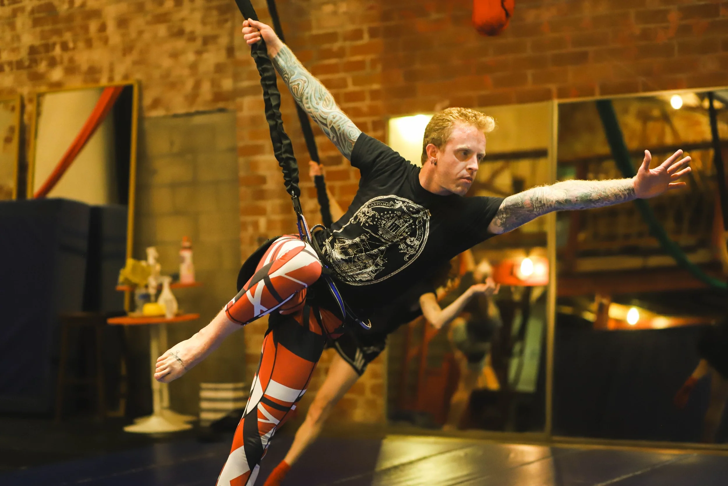 A man with tattoos on his arms and curly hair is performing a stretching exercise in a gym, holding a suspension trainer with his right hand while extending his left arm forward.