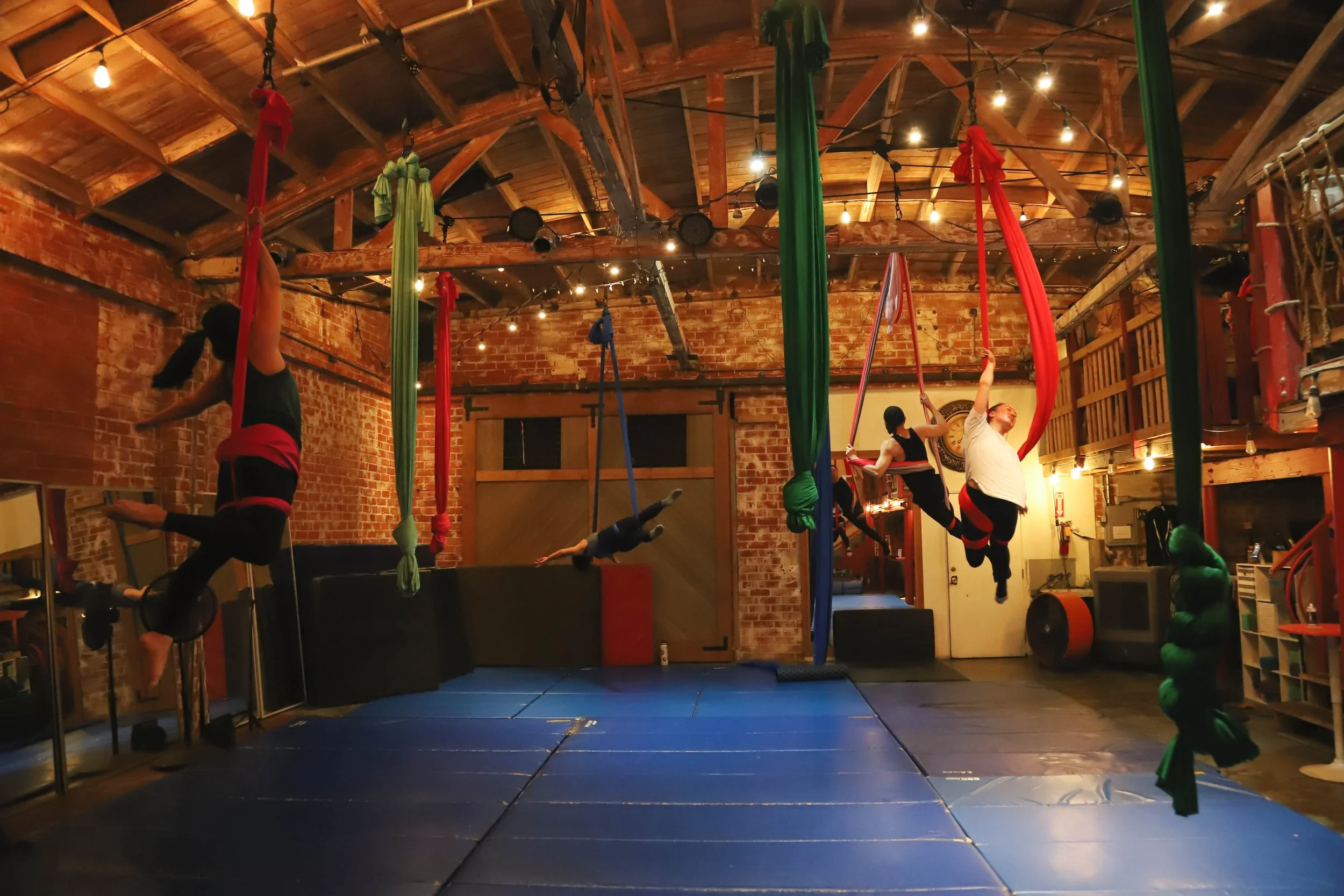 People practicing aerial silk and trapeze in a rustic indoor gym with brick walls and wooden ceiling, decorated with string lights.