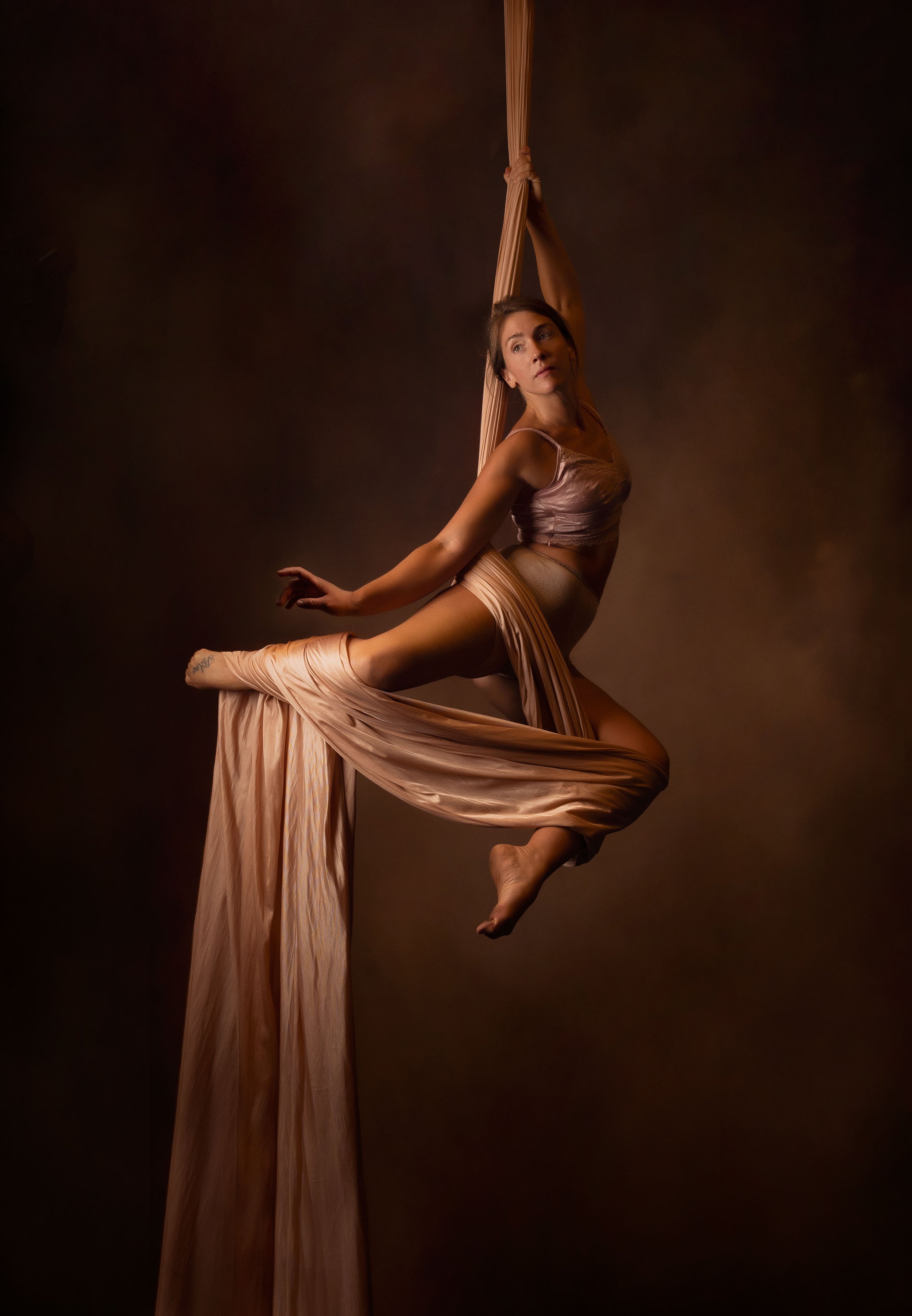 A woman performing an aerial dance or acrobatics with beige silk fabric, suspended in mid-air against a dark background.