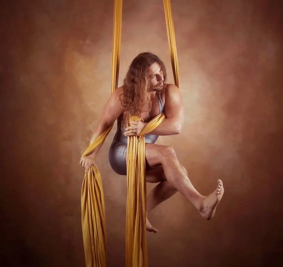 A man with curly hair on a yellow aerial silk, against a brown background.