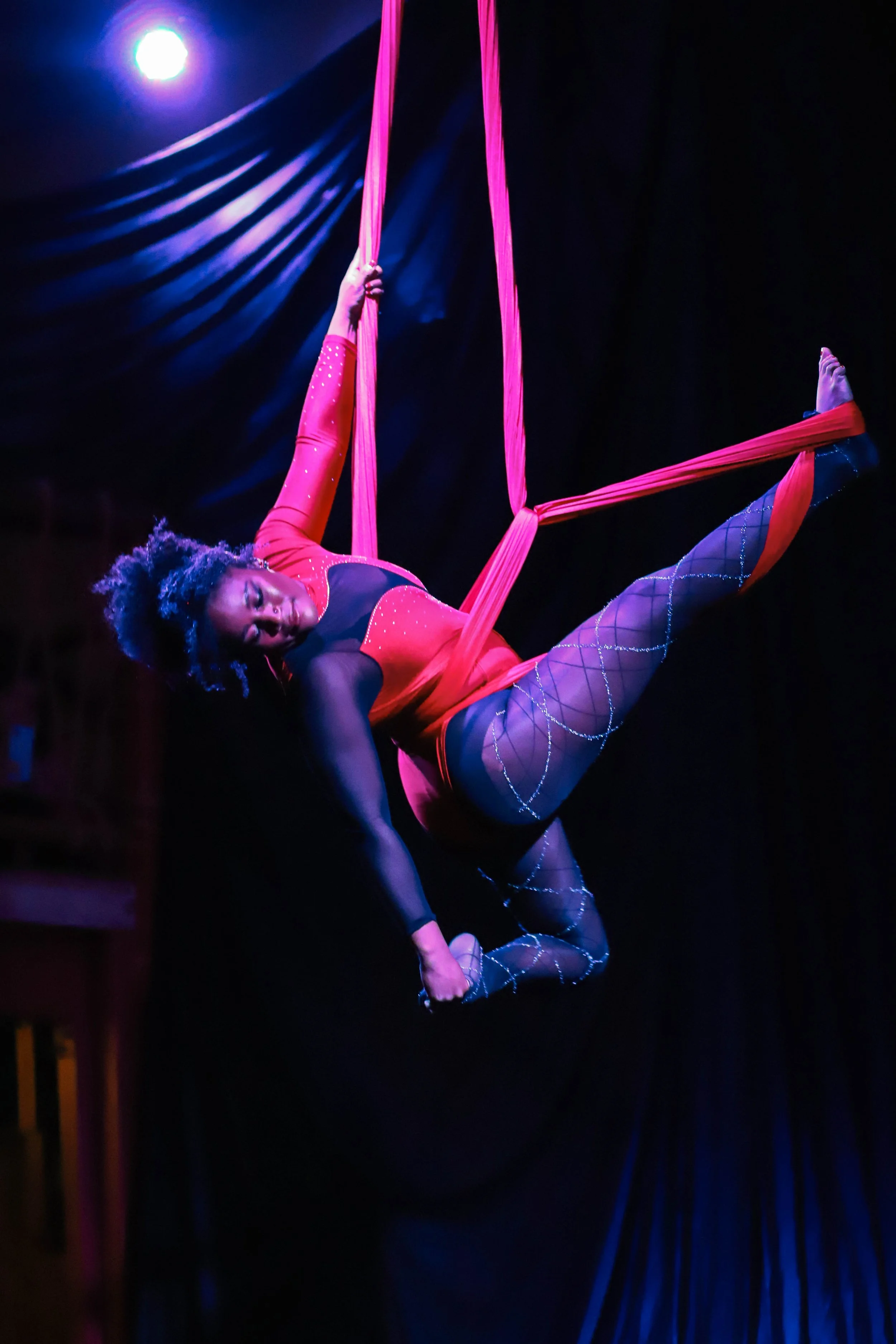 A female aerial silk performer suspended in the air, holding onto pink silks with one hand and extending her leg, wearing a red and black costume with fishnet tights, under stage lighting against a dark background.