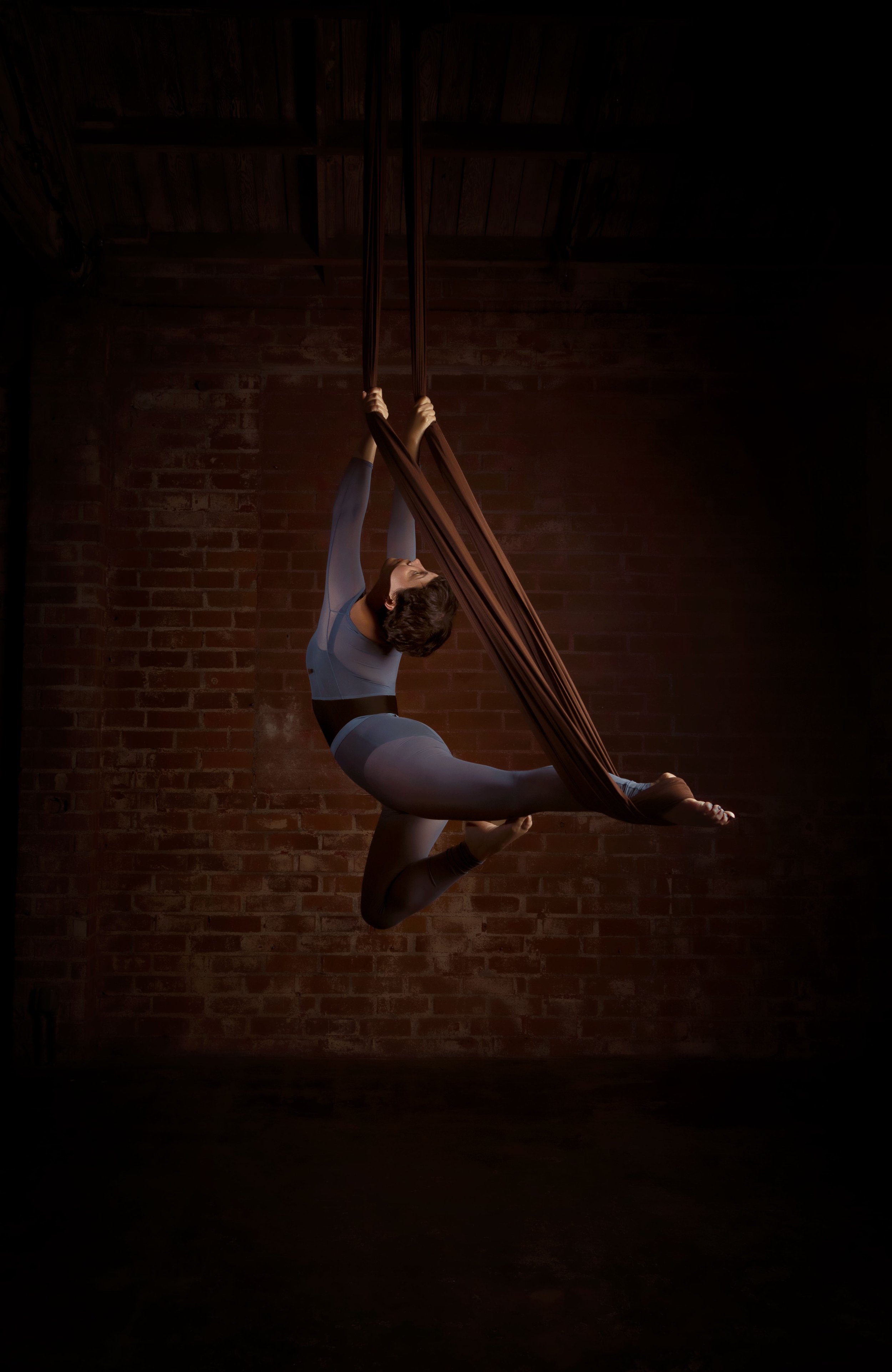 A female aerial silk performer hangs upside down on brown silk fabrics against a red brick wall, in a dark studio environment with spotlight highlighting her.