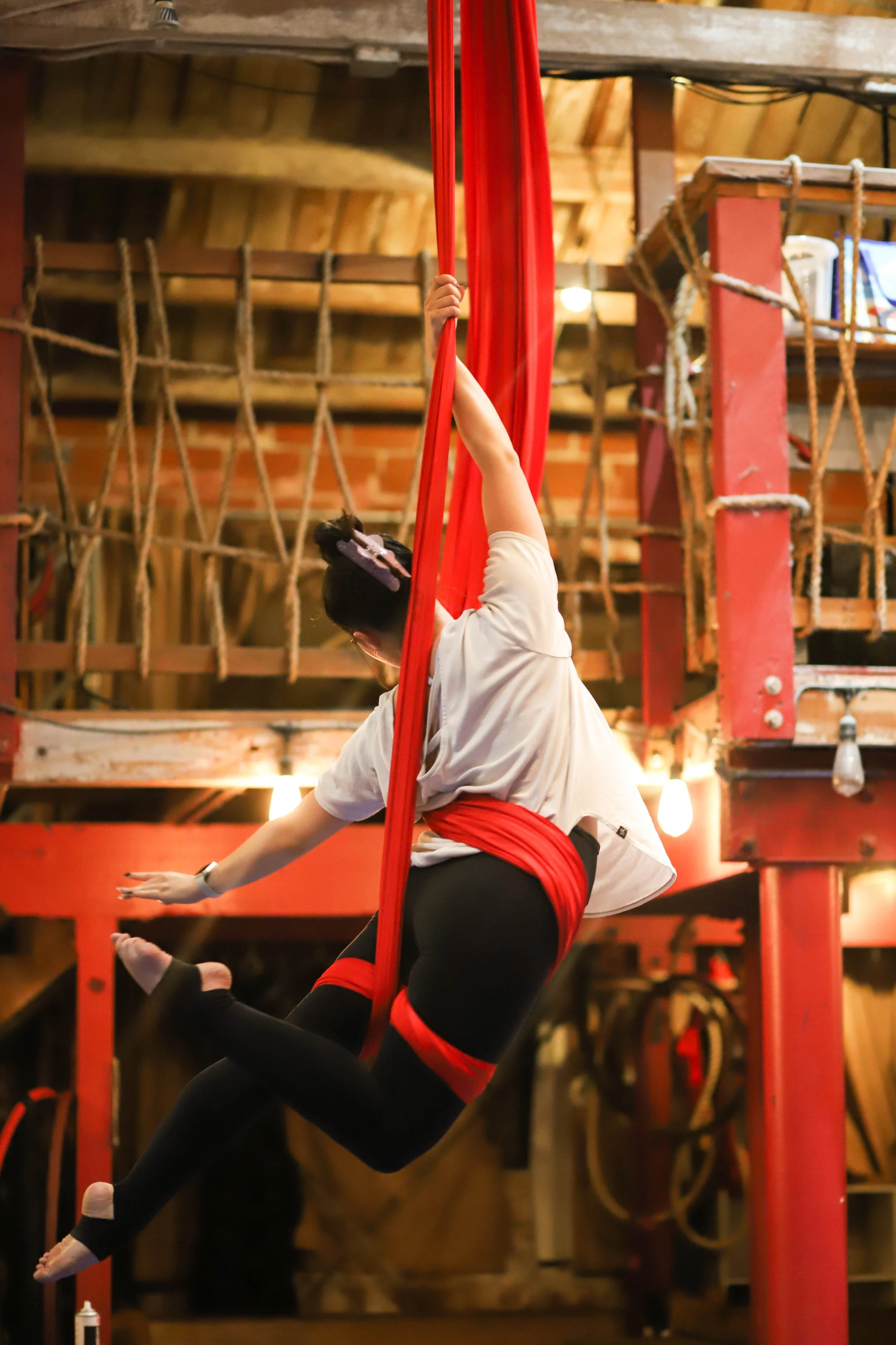 A person performing aerial silk acrobatics on red silks inside a room with wooden beams and ropes, with another person visible beneath them.
