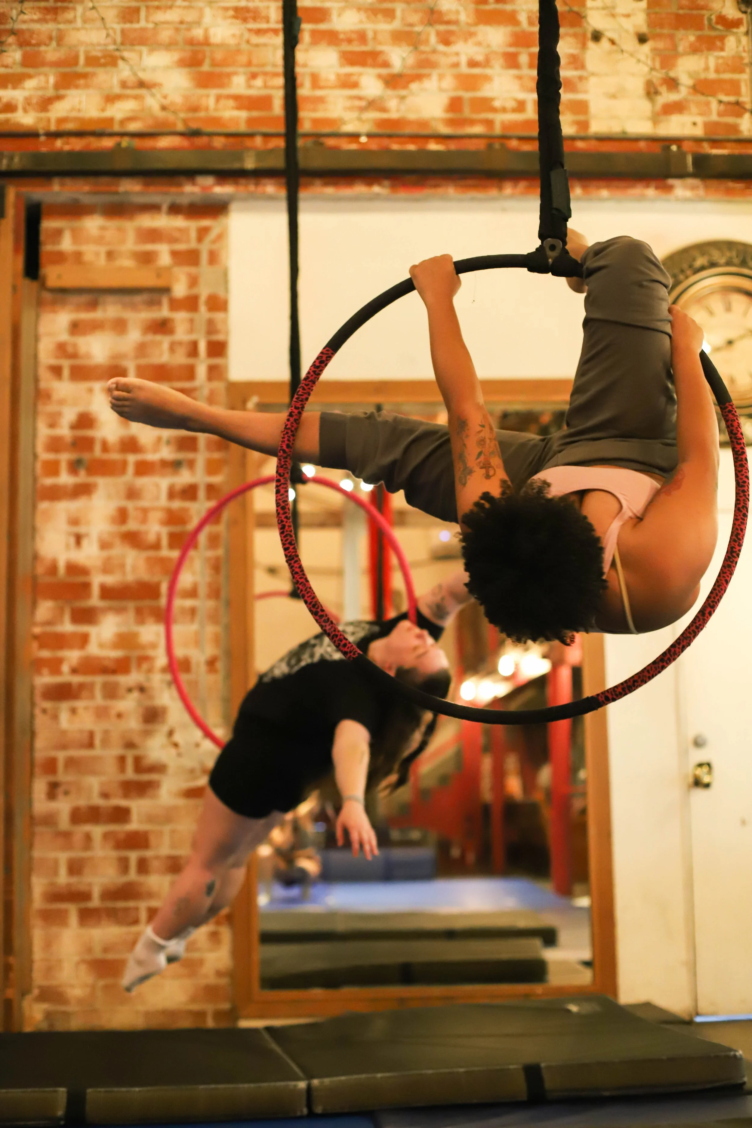 Person hanging upside down inside a red and black hoop in a gym with exposed brick wall.