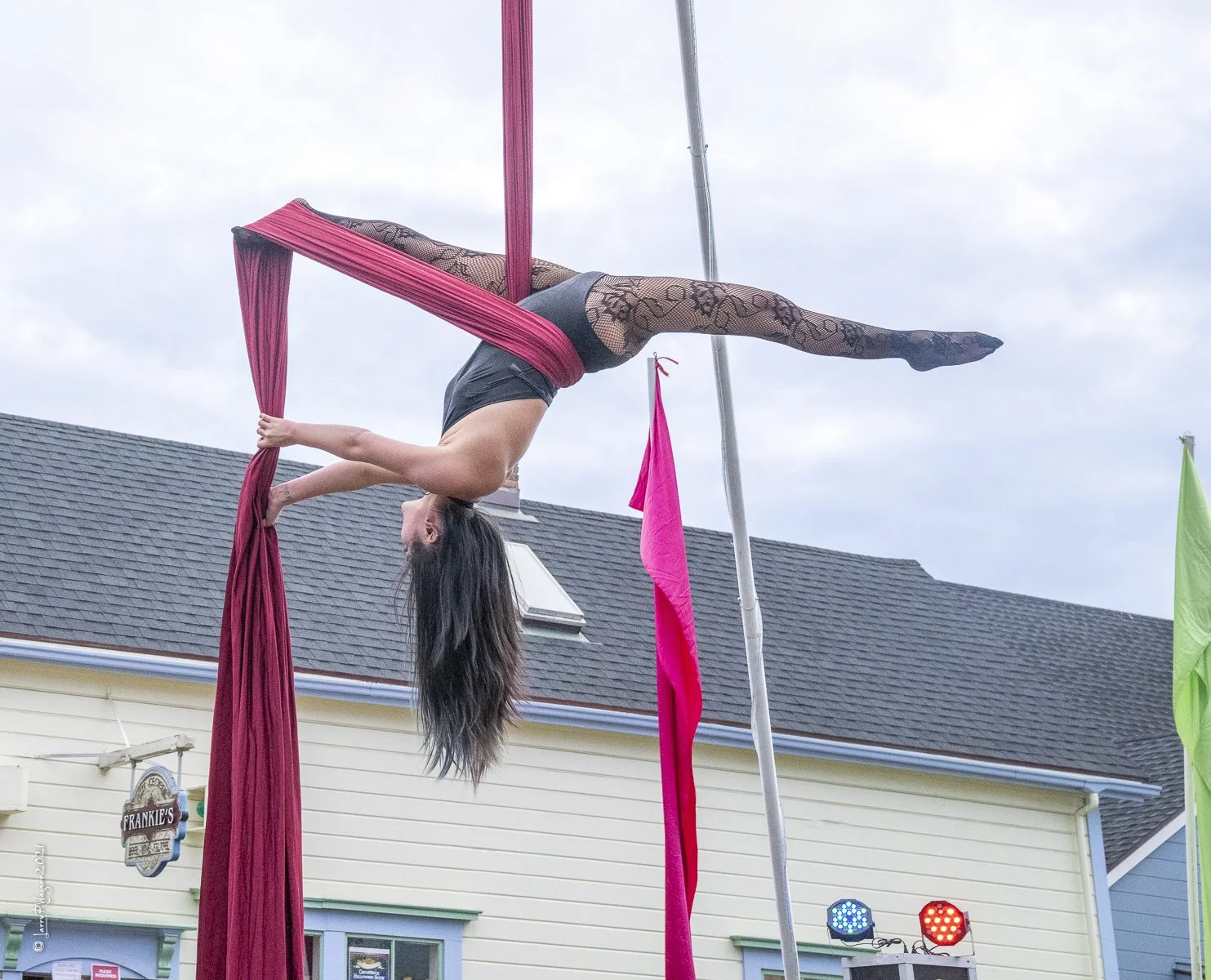 Aerial silk performer hanging upside down from pink silks outdoors, performing an acrobatic pose with the body extended horizontally and head downward.
