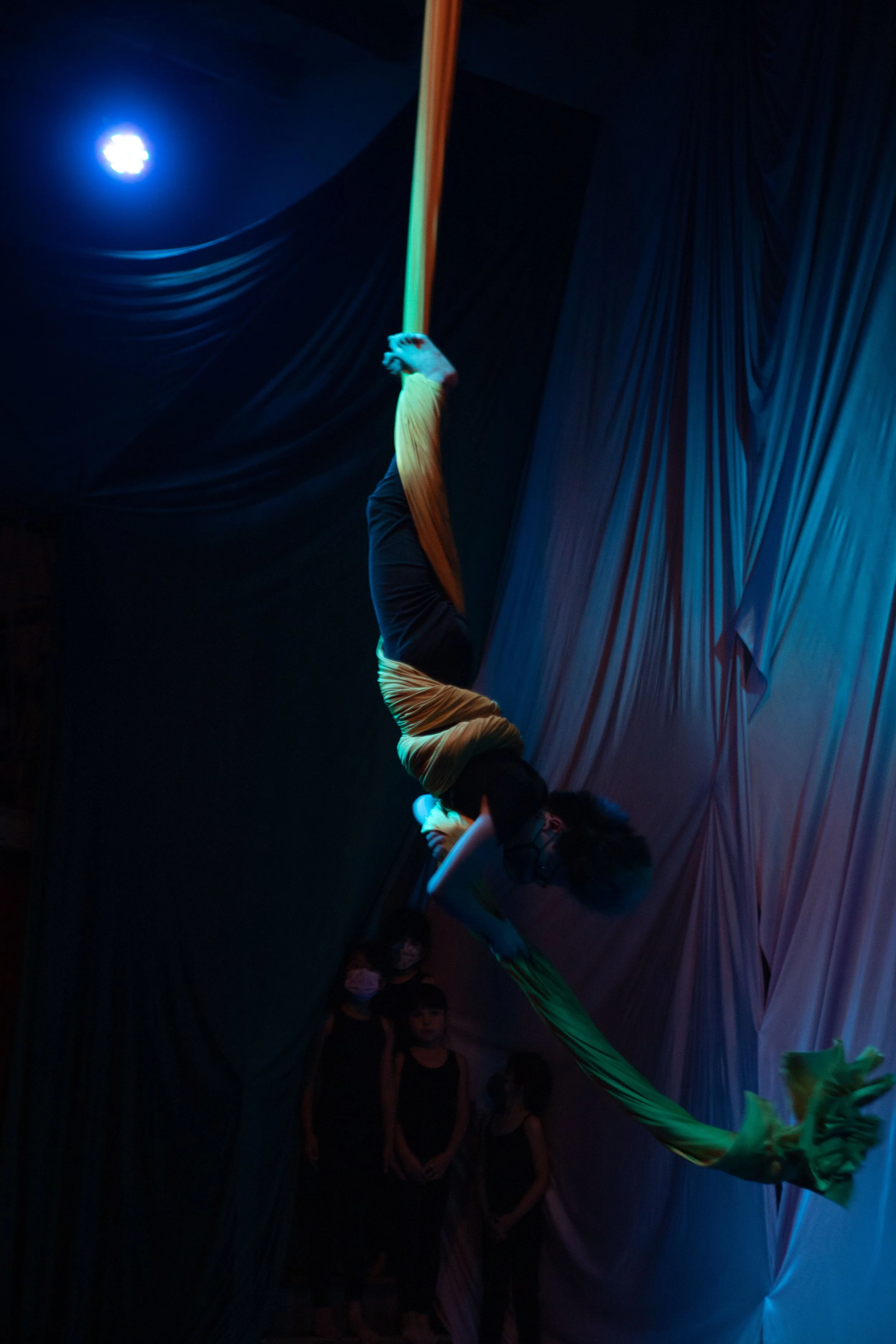 A performer hanging upside down with legs wrapped around a yellow fabric, surrounded by a dark stage with blue and purple lighting, while an audience watches.