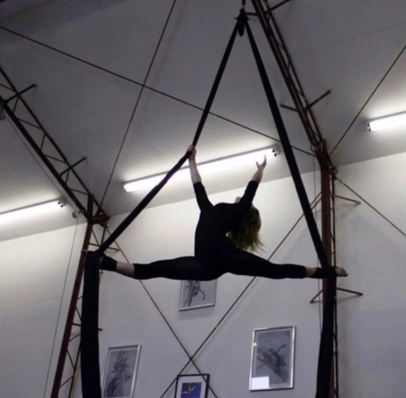 A female aerialist performing a split on aerial silks inside a gym or training facility with framed pictures on the wall.
