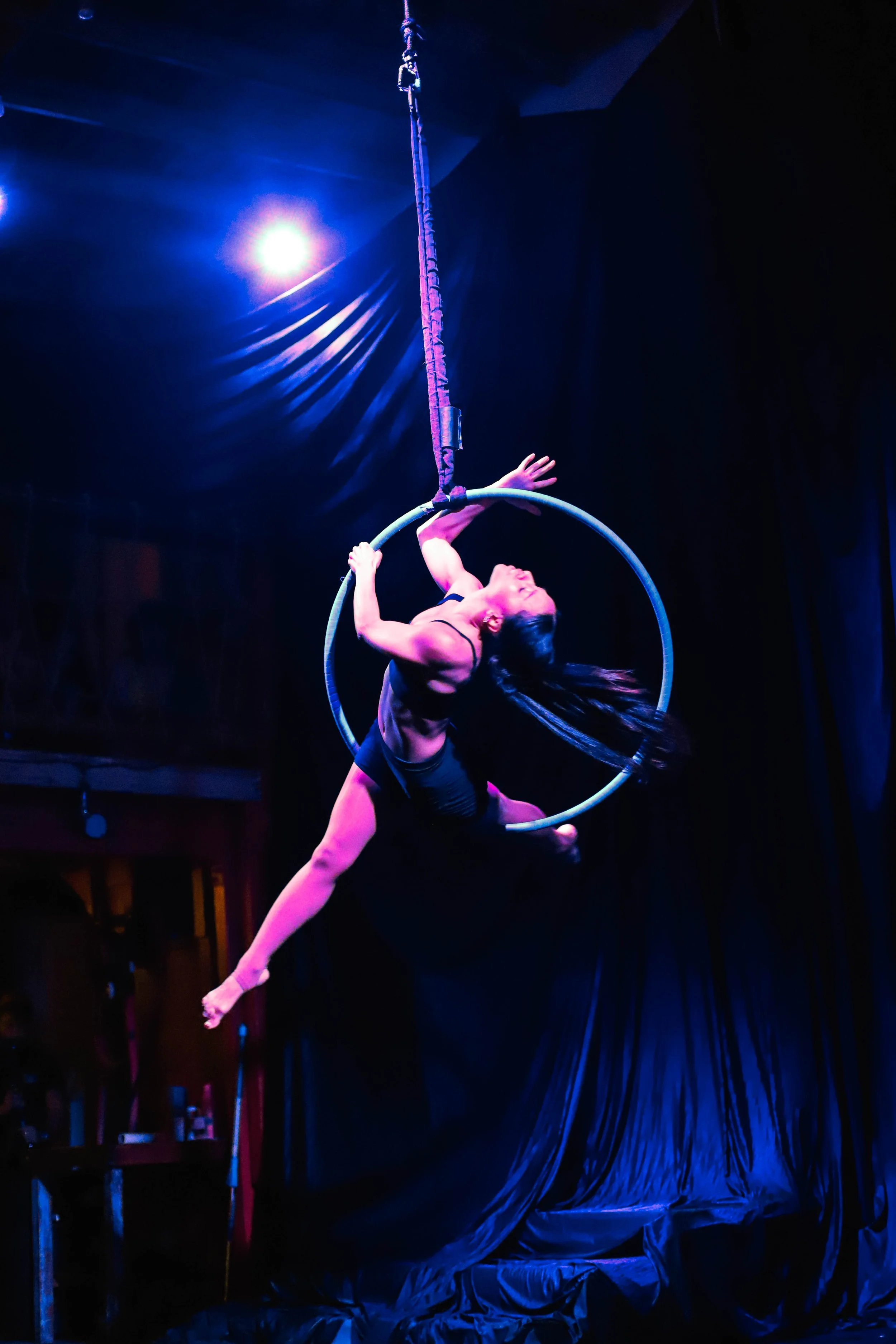 Circus performer performing aerial act on a hoop and hanging from a rope, illuminated by stage lights in a black background.