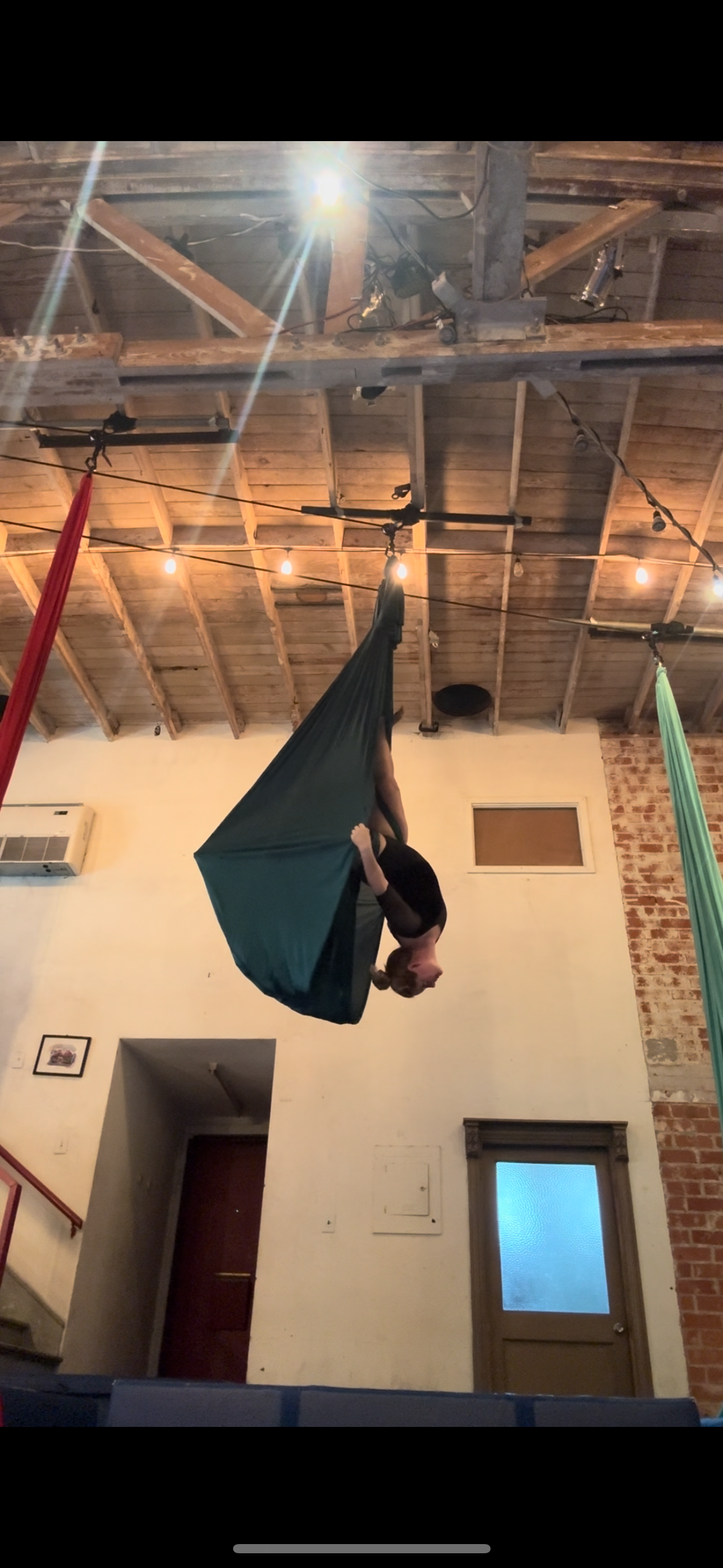 A woman practicing aerial silk acrobatics in a loft-style room with exposed wood beams and string lights, hanging upside down from a green fabric.