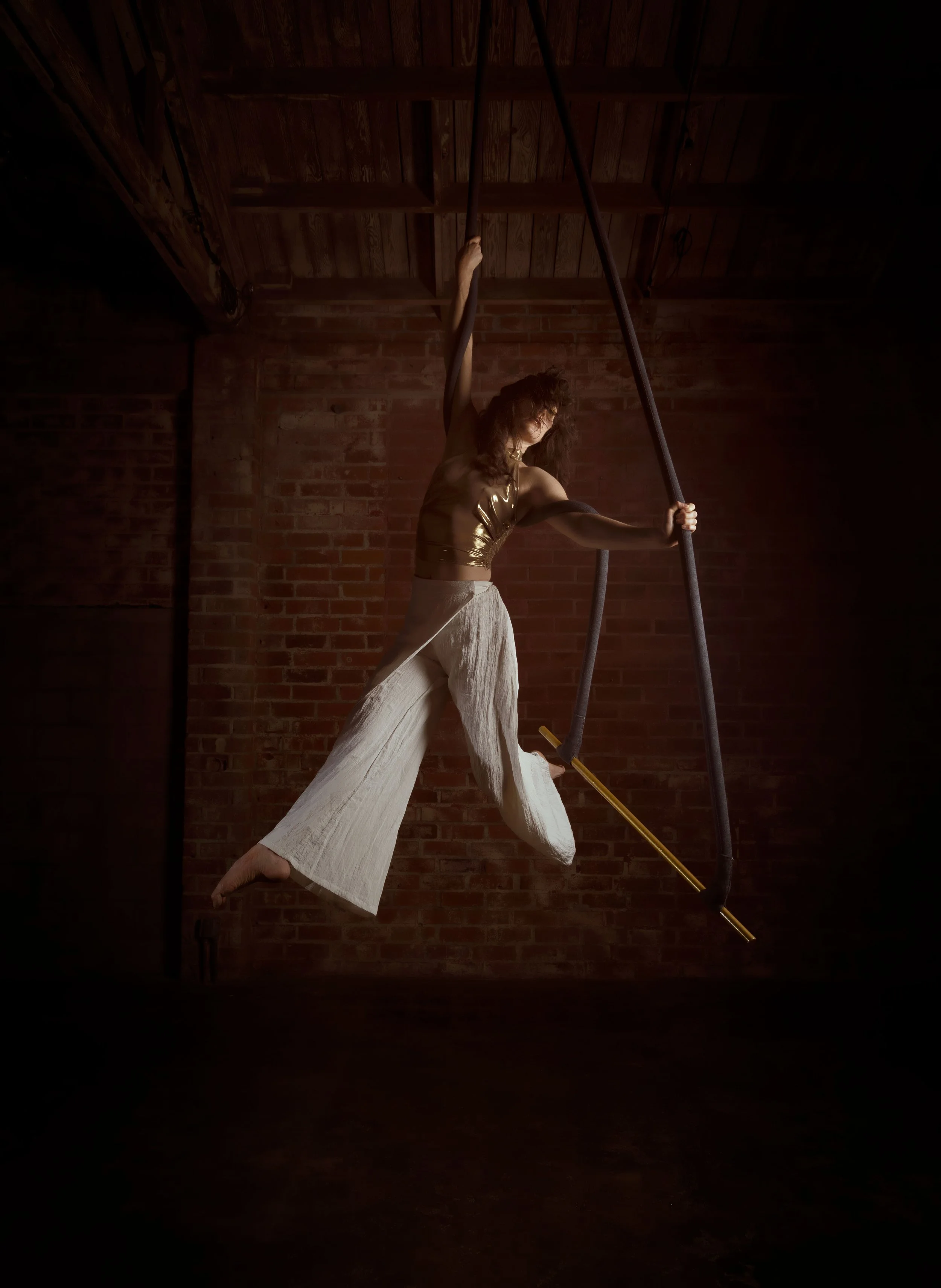 A woman with curly hair wearing a shiny gold top and loose white pants hanging from a trapeze or aerial silk, performing an aerial dance or gymnastics move in a dimly lit room with brick walls and wooden ceiling.