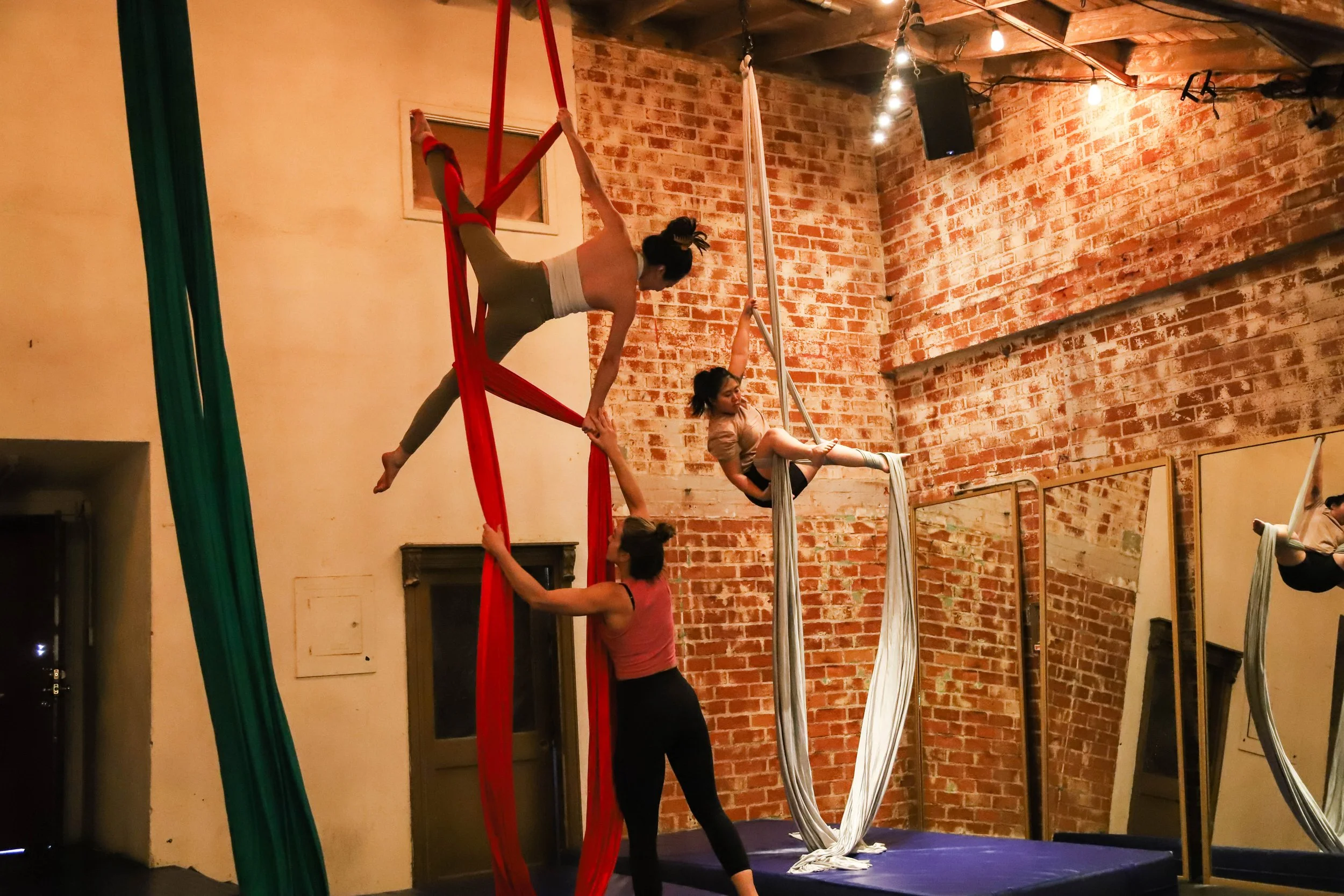 Two women practicing aerial silk acrobatics on red silks, with one woman in mid-air performing an upside-down move while another woman assists her. The scene takes place in a studio with brick walls and warm lighting.