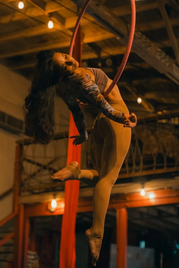 A female aerial performer hanging from a red hoop in a dimly lit indoor space.