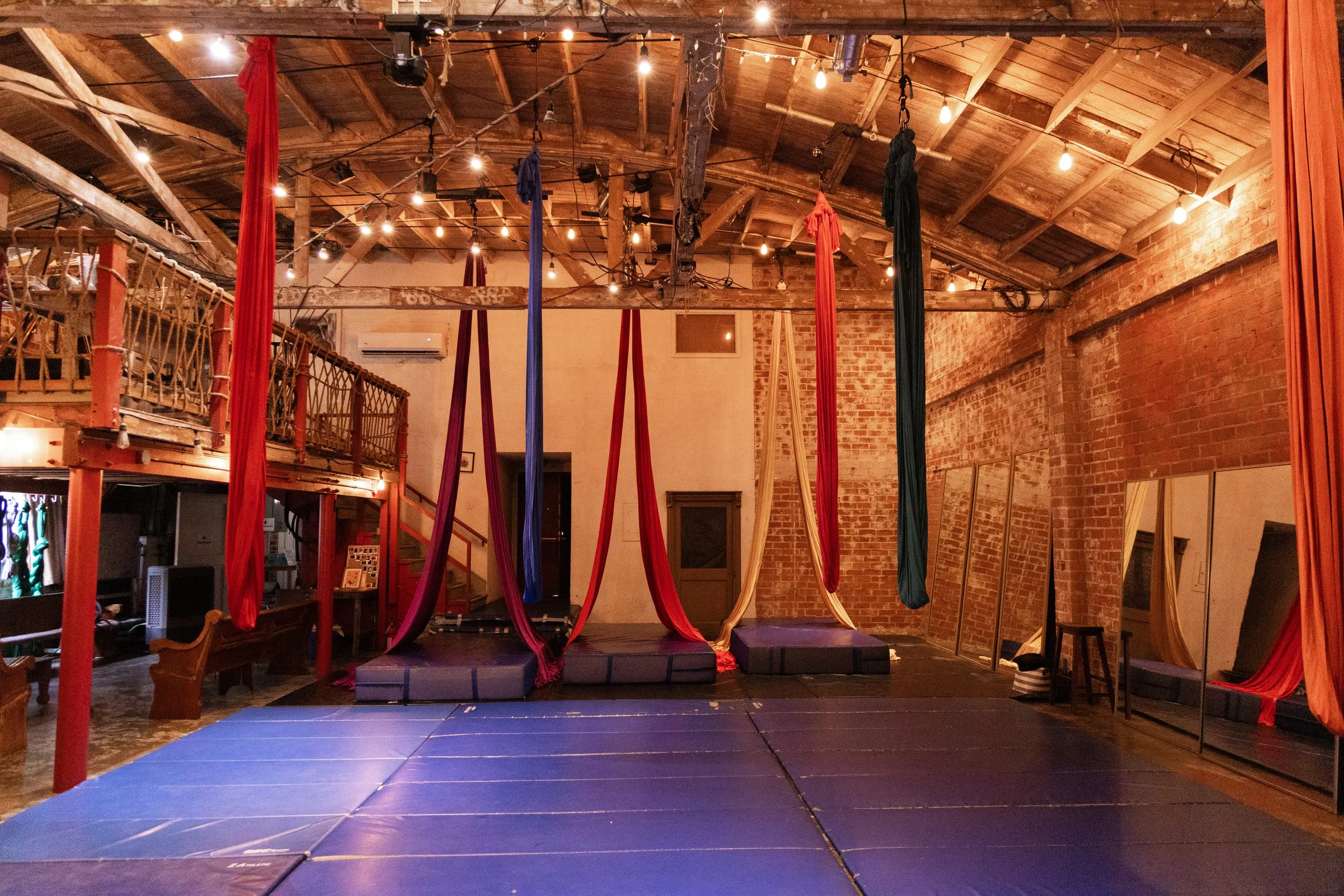 Indoor aerial arts space with fabric silks hanging from the ceiling over a padded performance area, exposed brick walls, hanging light bulbs, and a loft area to the left.