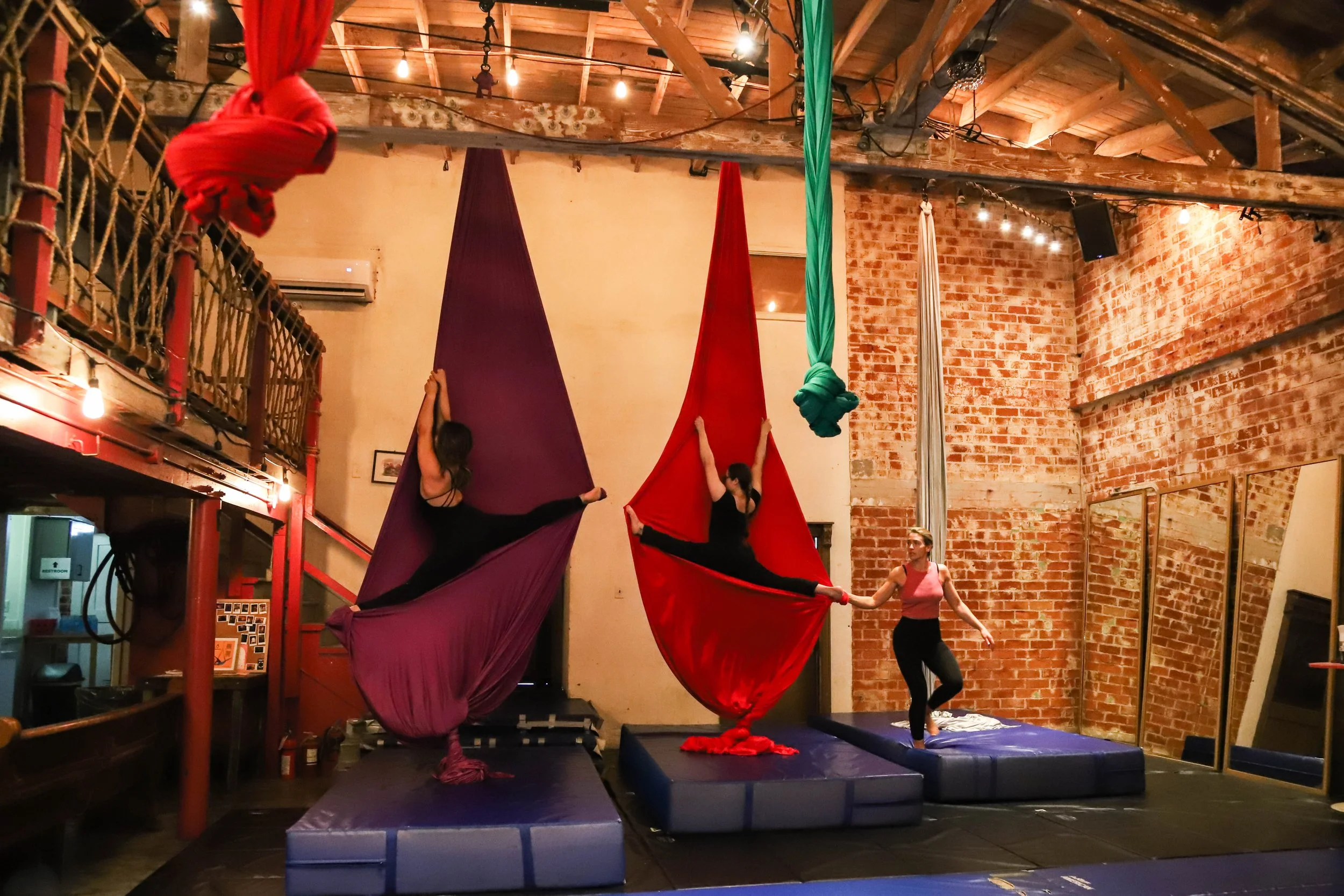 Two female aerial silk performers practicing in a studio with exposed brick walls and wooden ceiling beams, using red and purple silks, with mats for safety underneath.