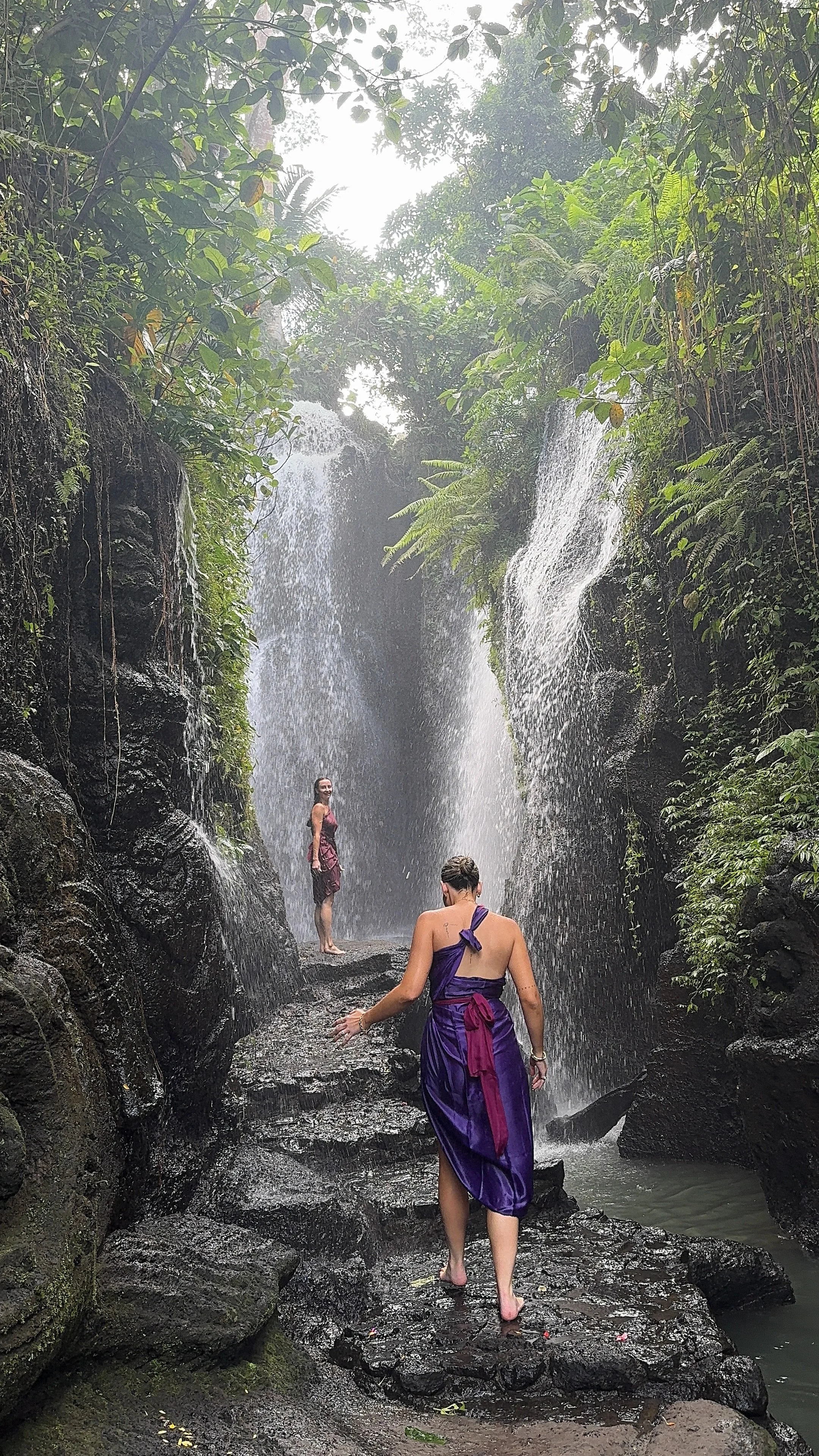 Two people stand beneath a tall jungle waterfall surrounded by lush tropical vegetation.