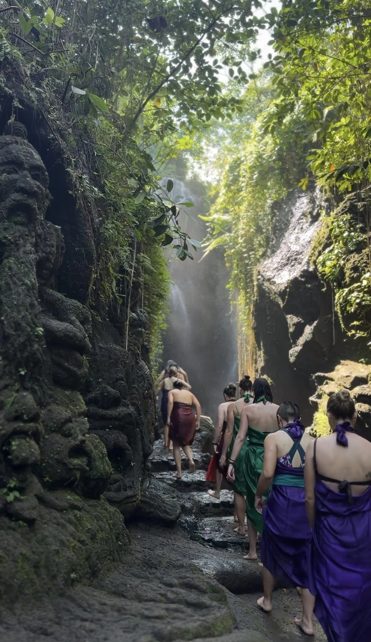 People in colorful sarongs walk through a lush, rocky gorge beneath a tropical waterfall.