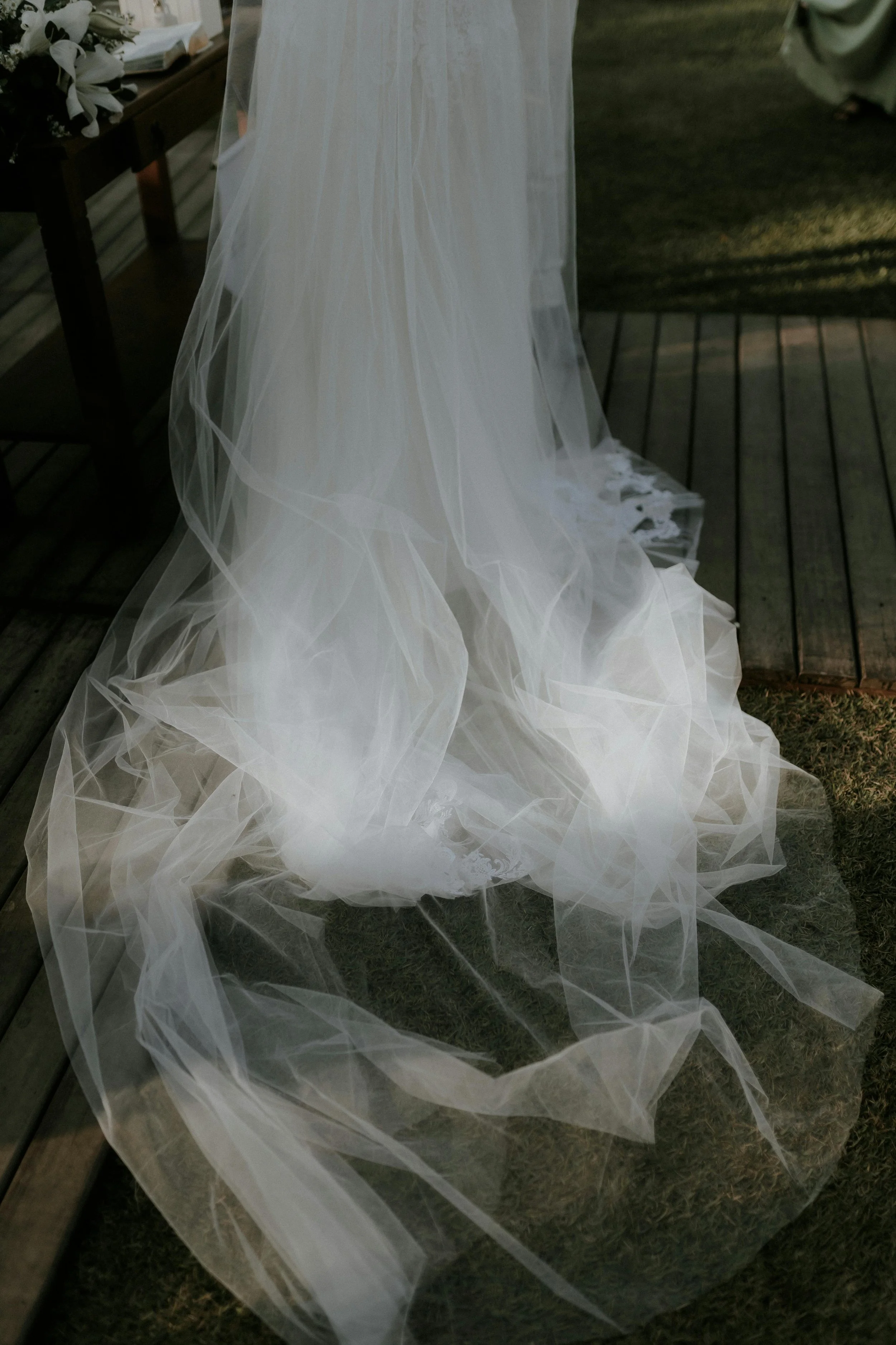 White wedding dress with a long sheer train draped over a wooden porch beside a bouquet of white flowers