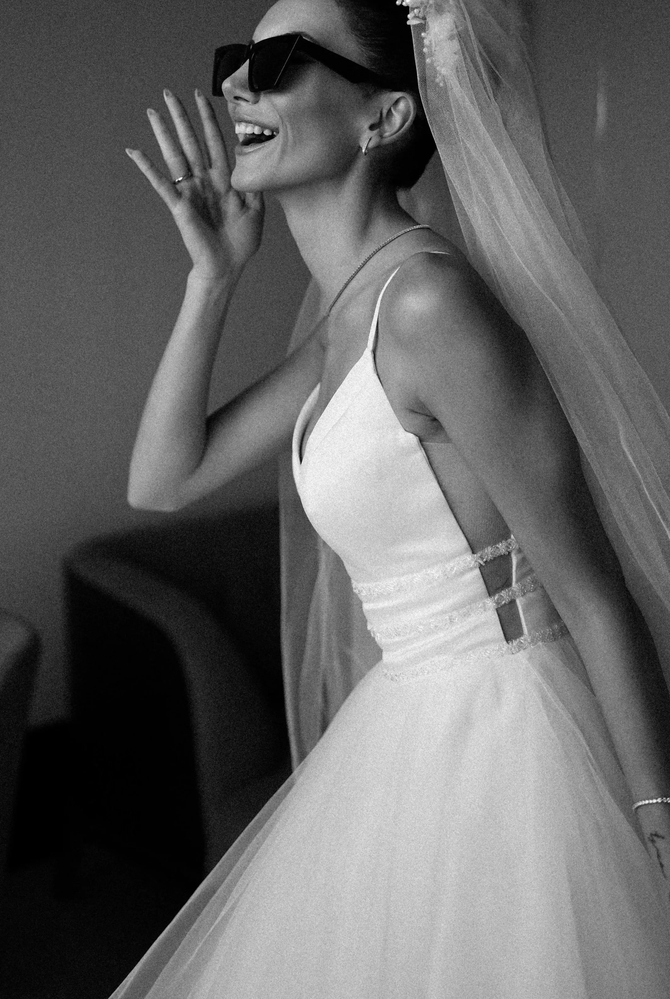 Black-and-white portrait of a smiling bride wearing oversized sunglasses, a veil, and a wedding dress.