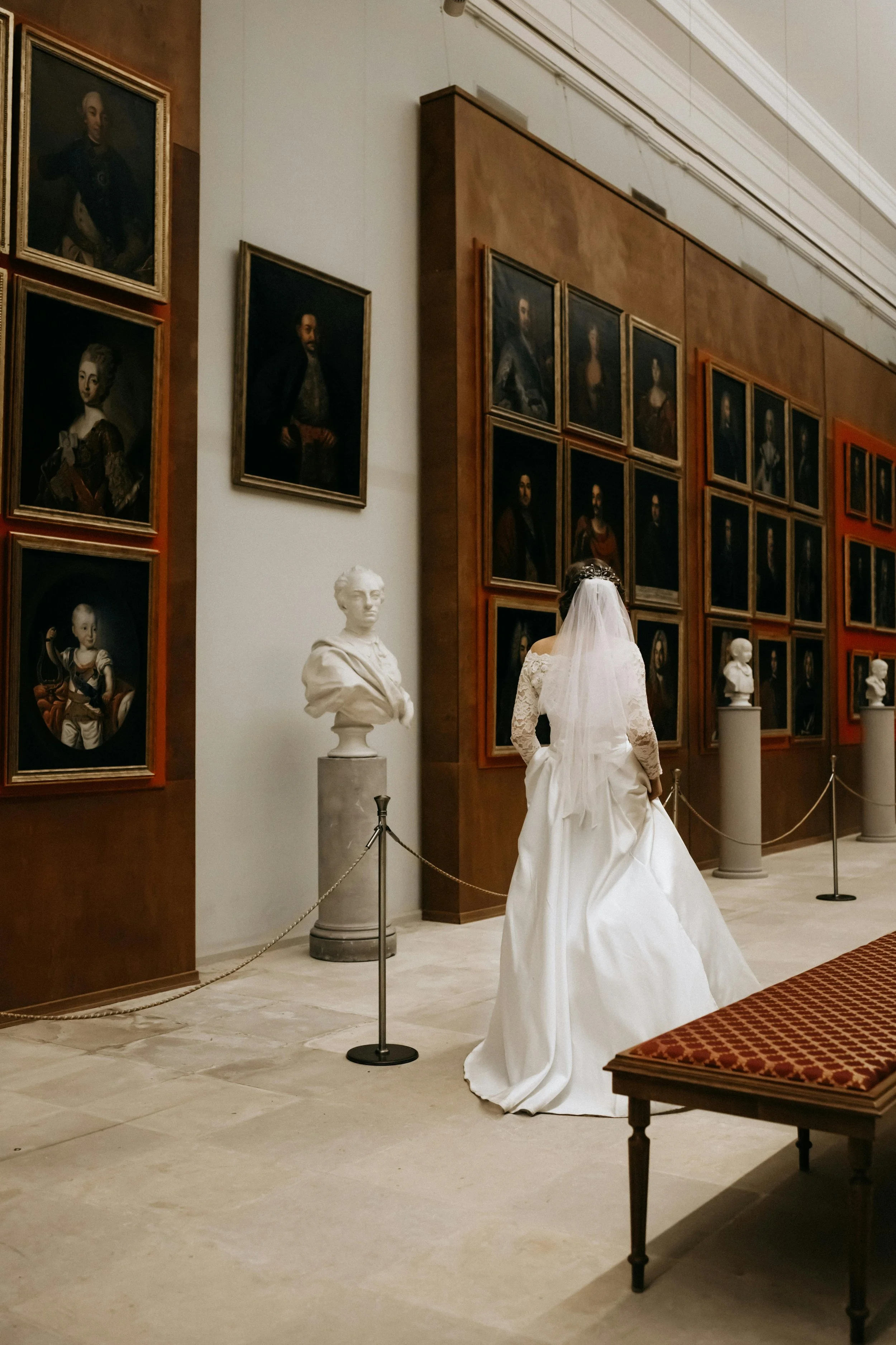 Bride in a white lace wedding gown and veil viewing framed historical portraits and marble busts in a museum gallery.