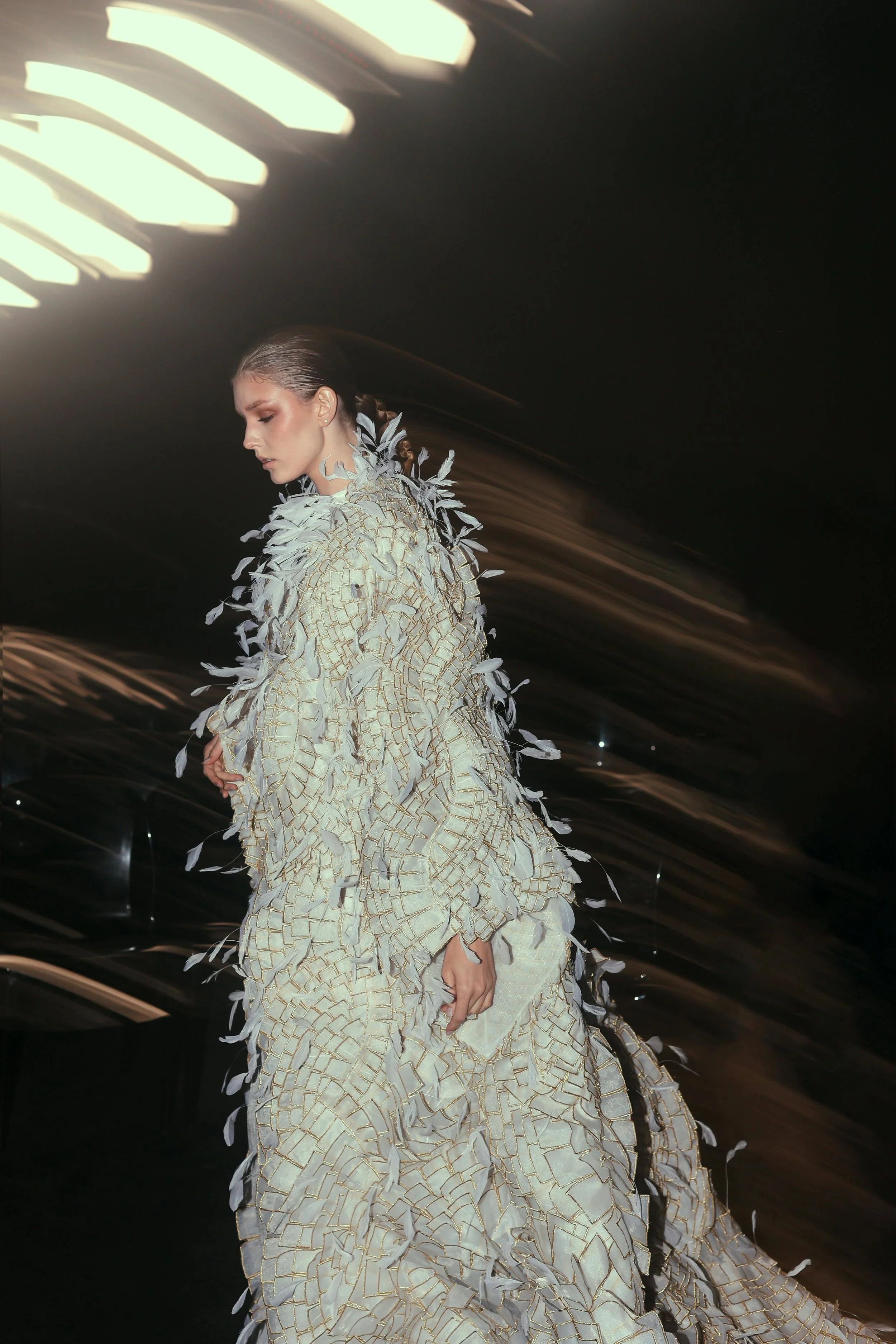 Woman in an ornate white and gold feathered gown posing beneath dramatic overhead lights.