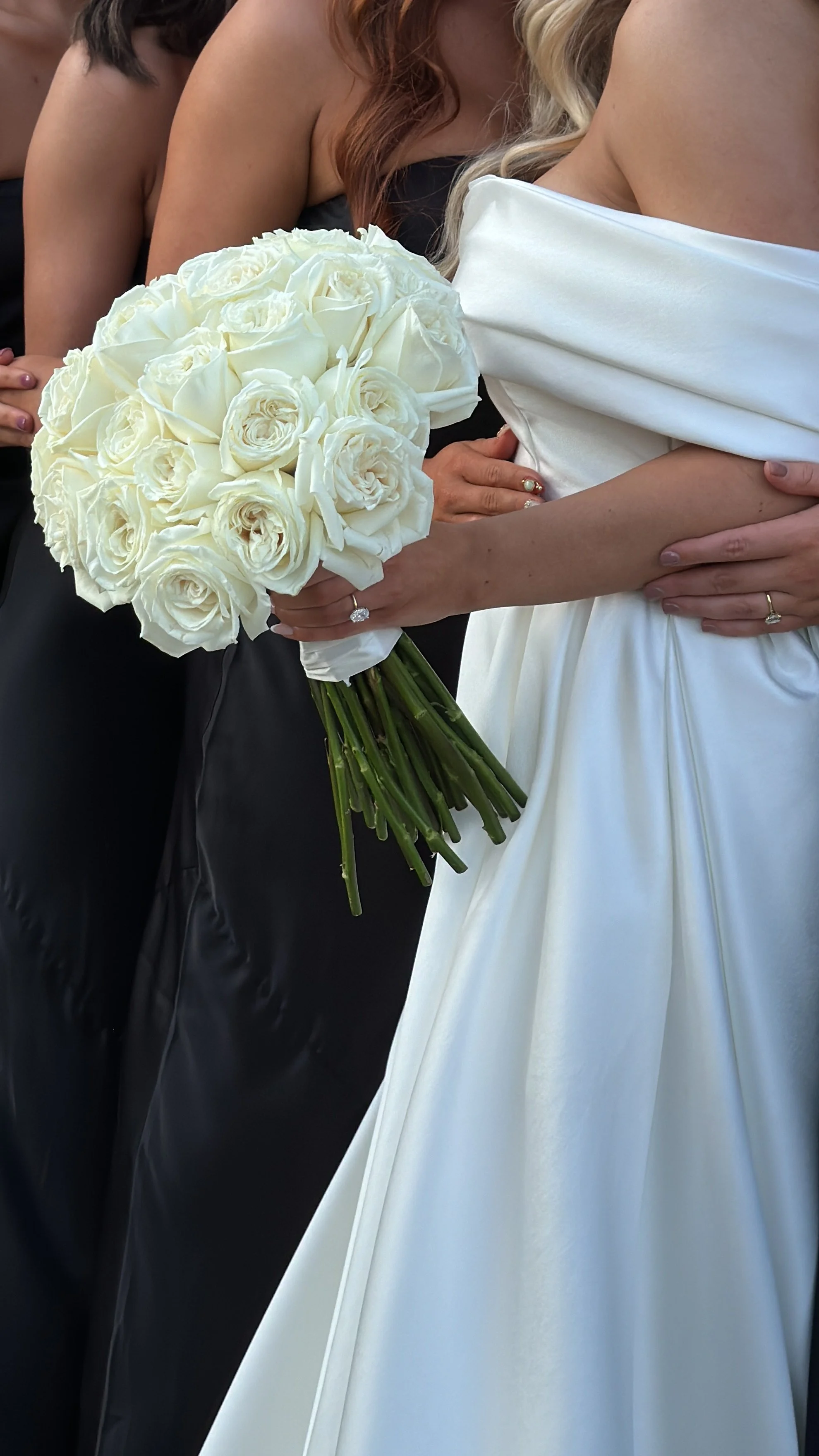 Bride in a white wedding dress holding a bouquet of white roses beside bridesmaids in black dresses