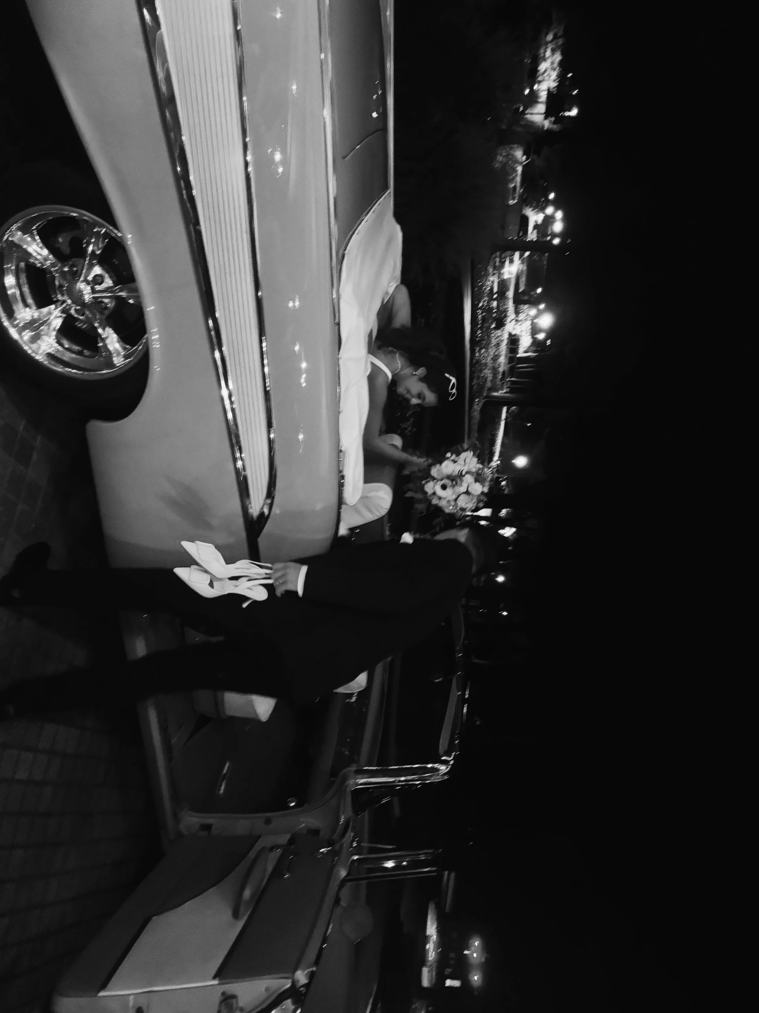 Black-and-white photo of a bride and groom embracing beside a decorated vintage car at night