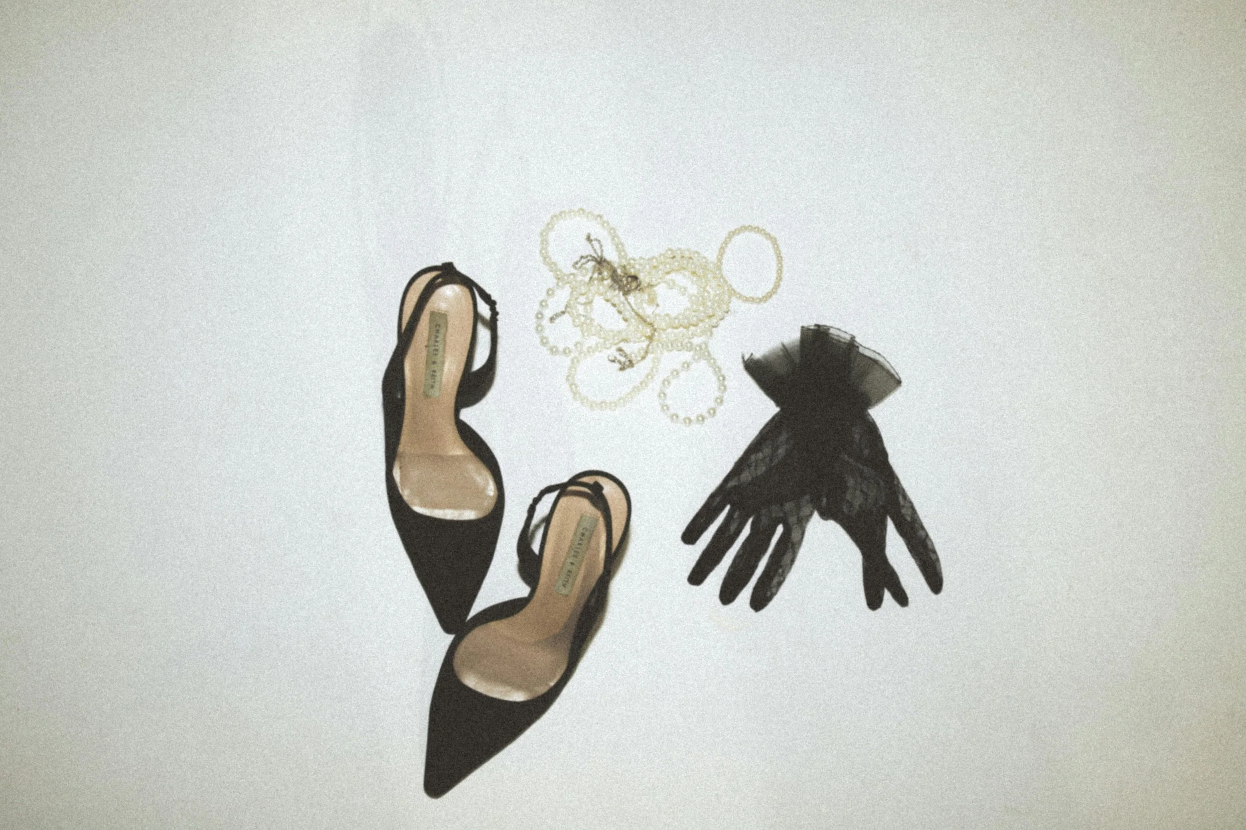 Black pointed-toe high heels, pearl necklace, and black lace gloves arranged on a white background