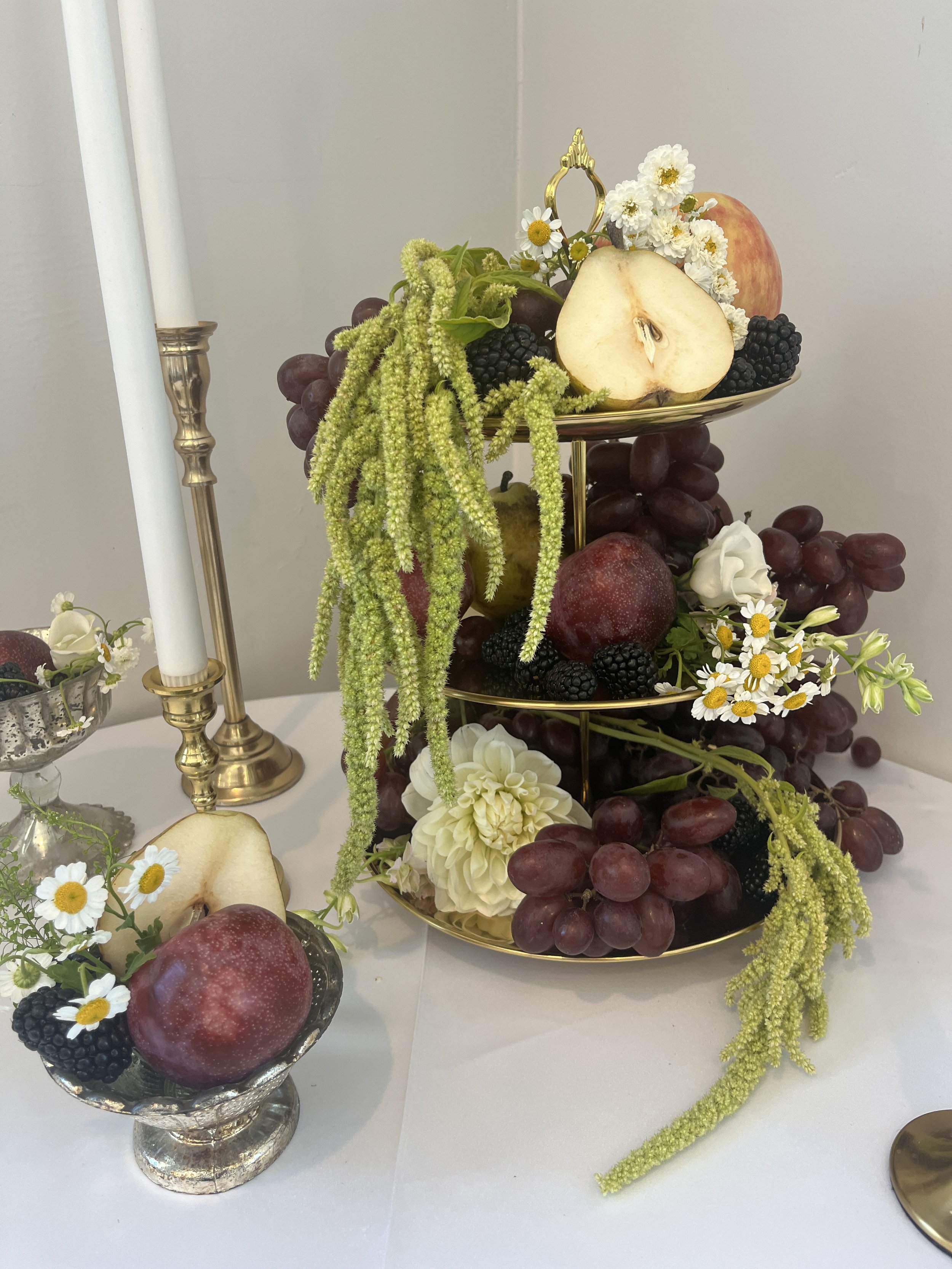 Elegant fruit and floral centerpiece with pears, apples, grapes, blackberries, white daisies, and candles on a tiered gold tray