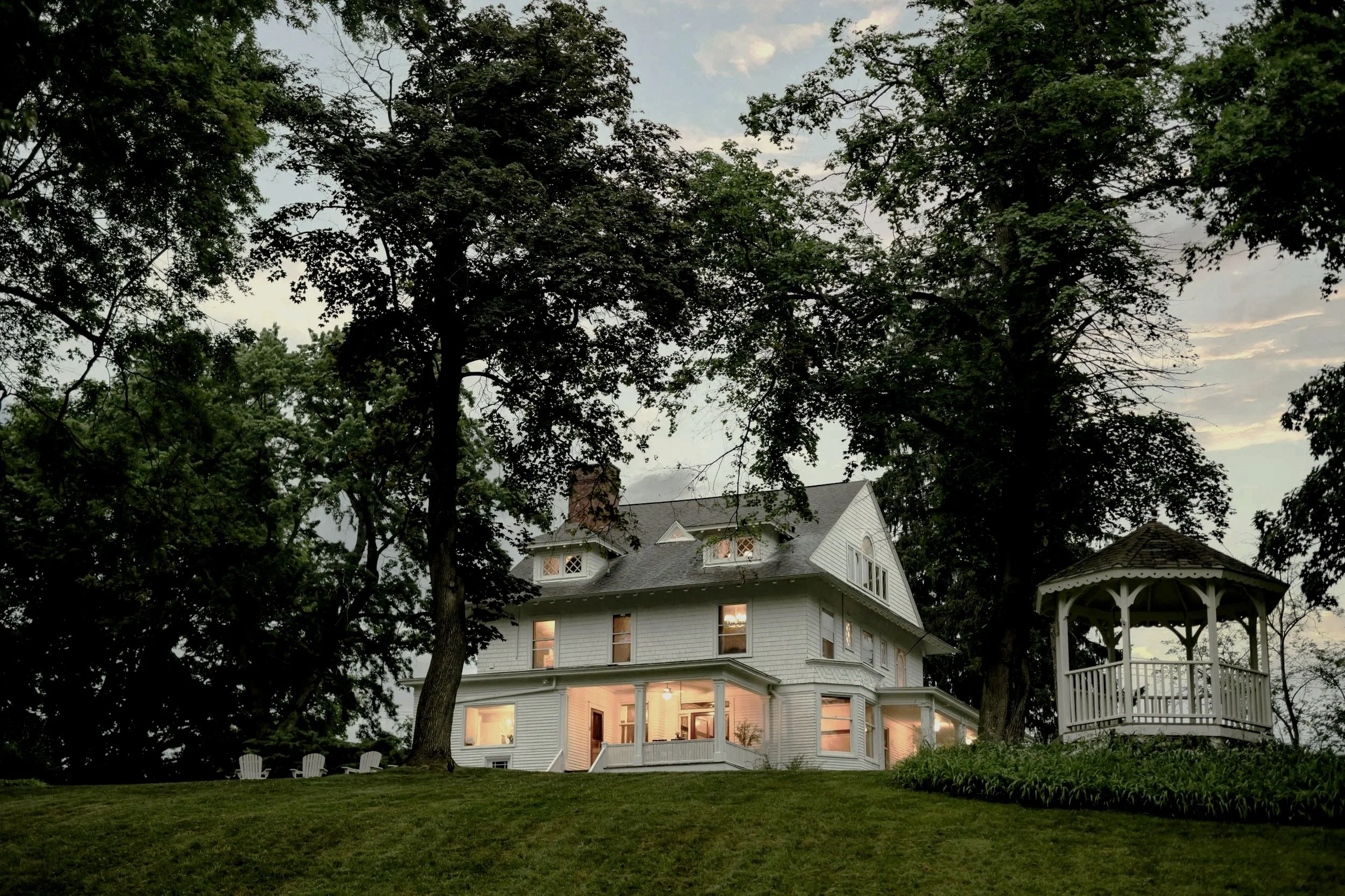Historic white Victorian house with lit windows and a gazebo surrounded by mature trees at dusk