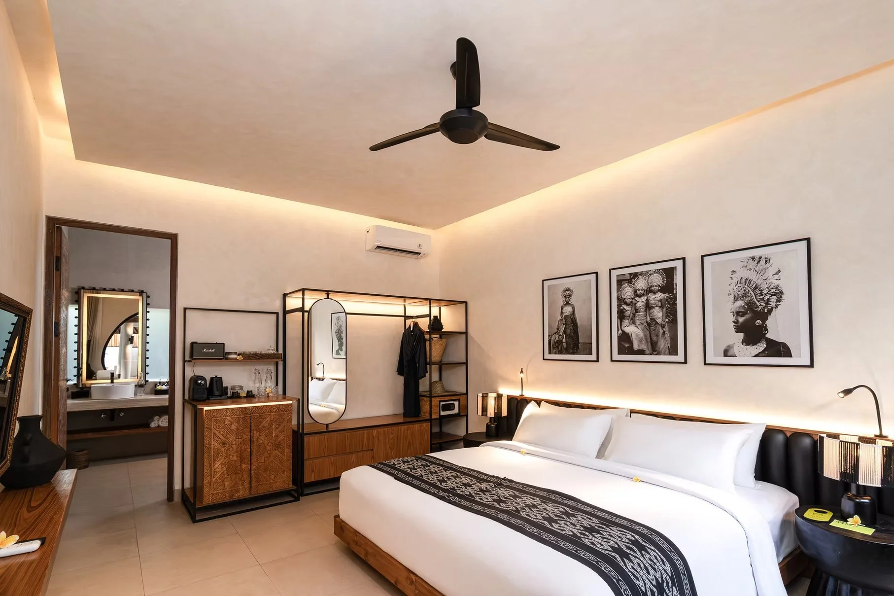 Modern tropical bedroom with a white bed, black patterned runner, ceiling fan, air conditioner, framed artwork, and wood furnishings
