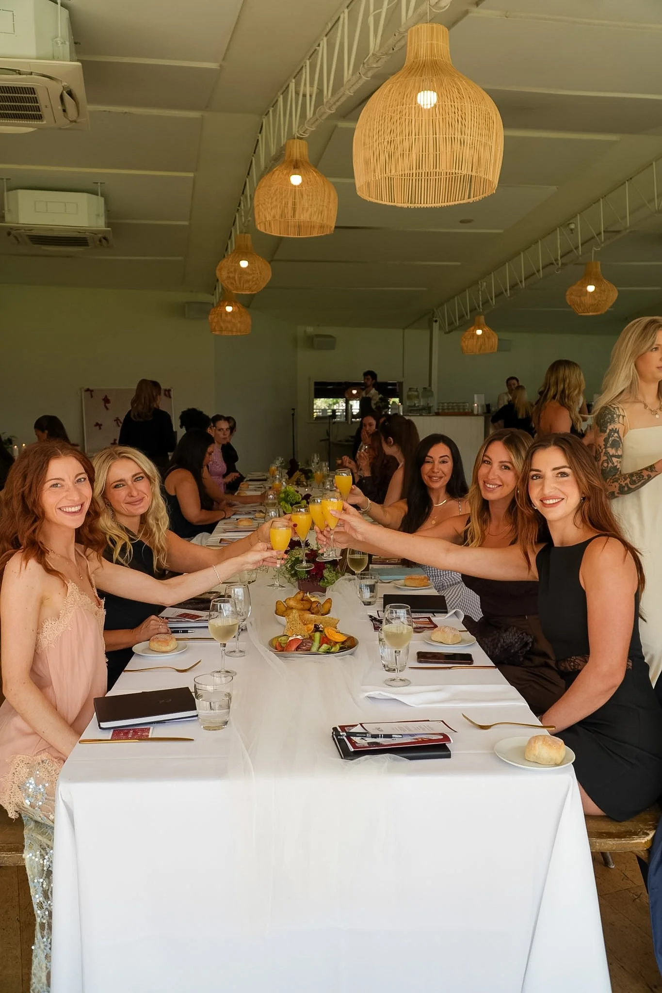 Group of women dining together and toasting with orange drinks at a restaurant table beneath woven pendant lights