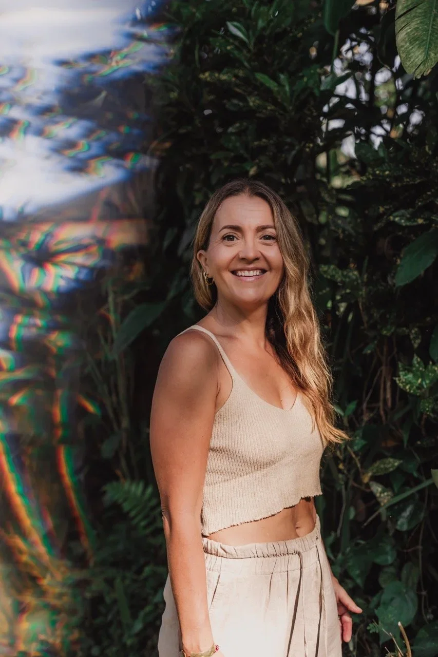 Smiling woman in a beige crop top and pants standing among lush tropical foliage with rainbow light reflections.