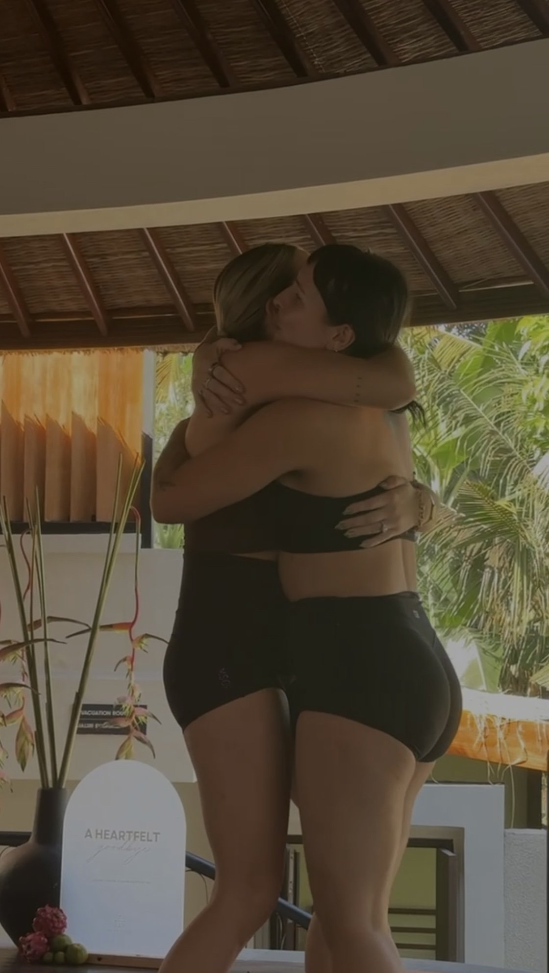 Two women embracing and kissing in a tropical outdoor setting beneath a thatched roof.